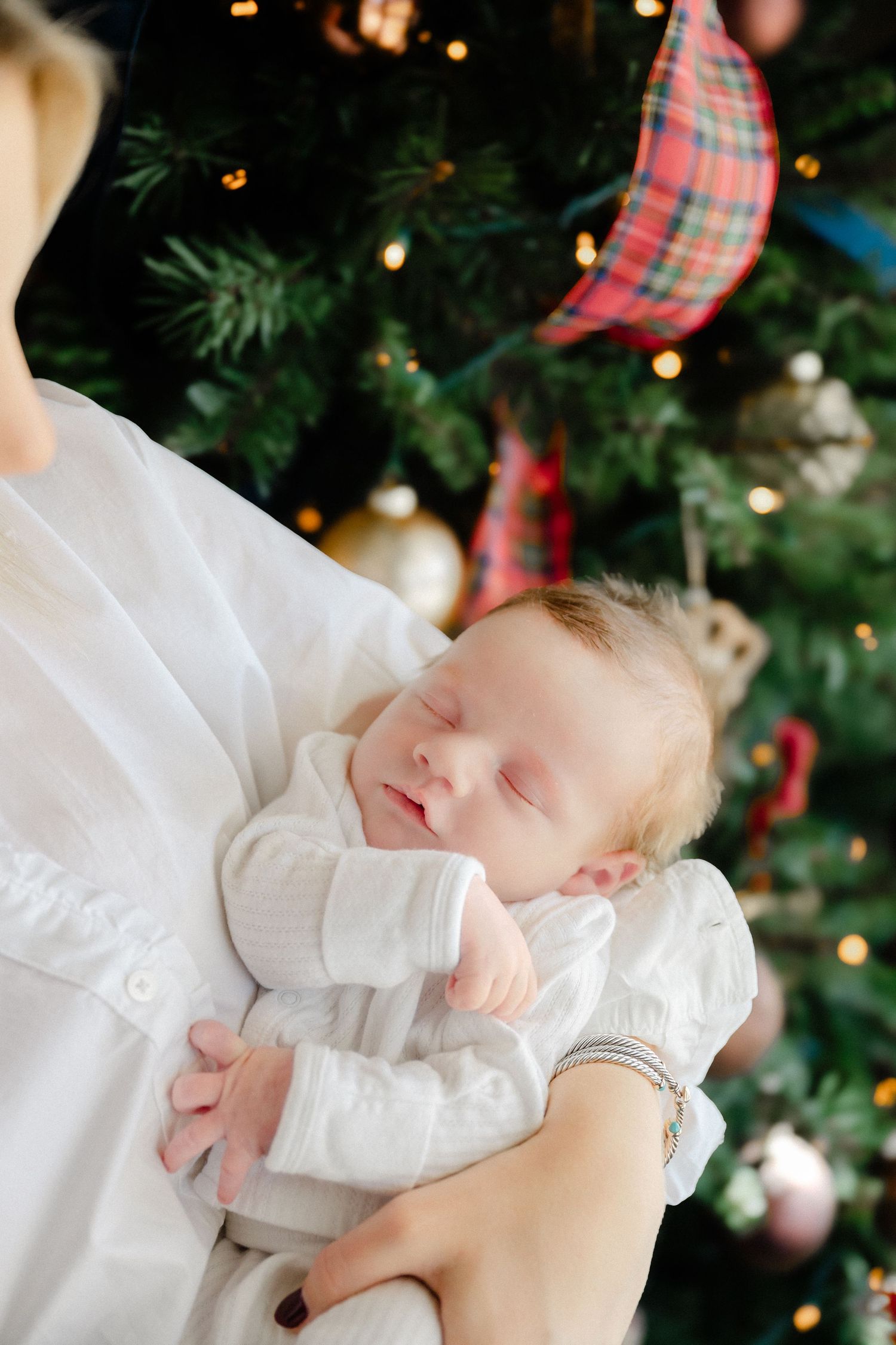 Sleeping newborn in white outfit against Christmas tree backdrop.