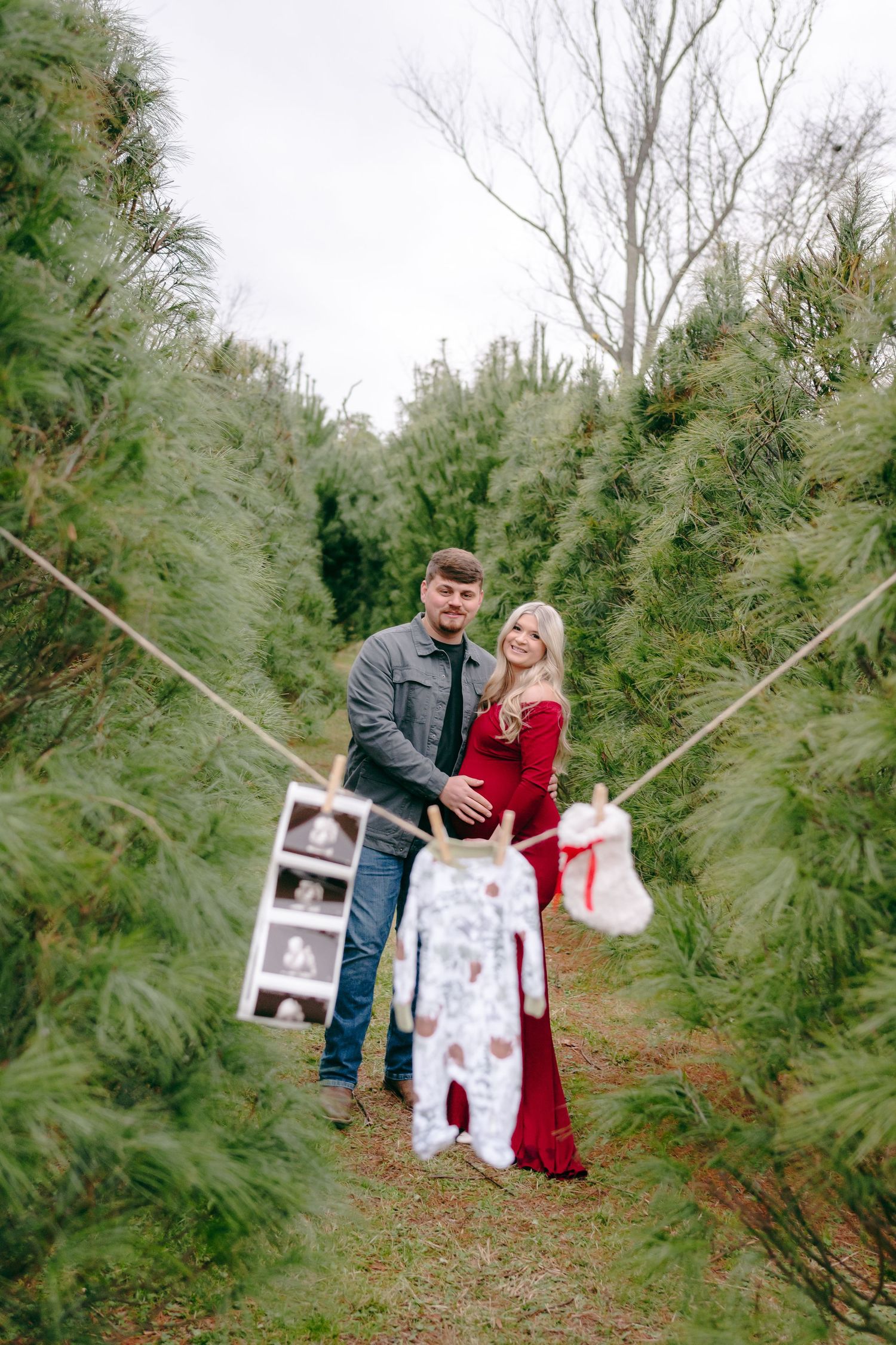 Couple displays ultrasound photos and Christmas stockings between rows of evergreen trees.