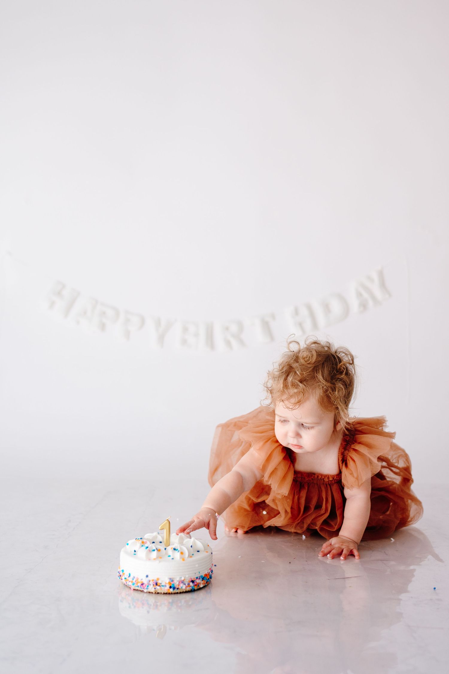 Baby in orange dress crawls towards birthday cake during cake smash photo session.