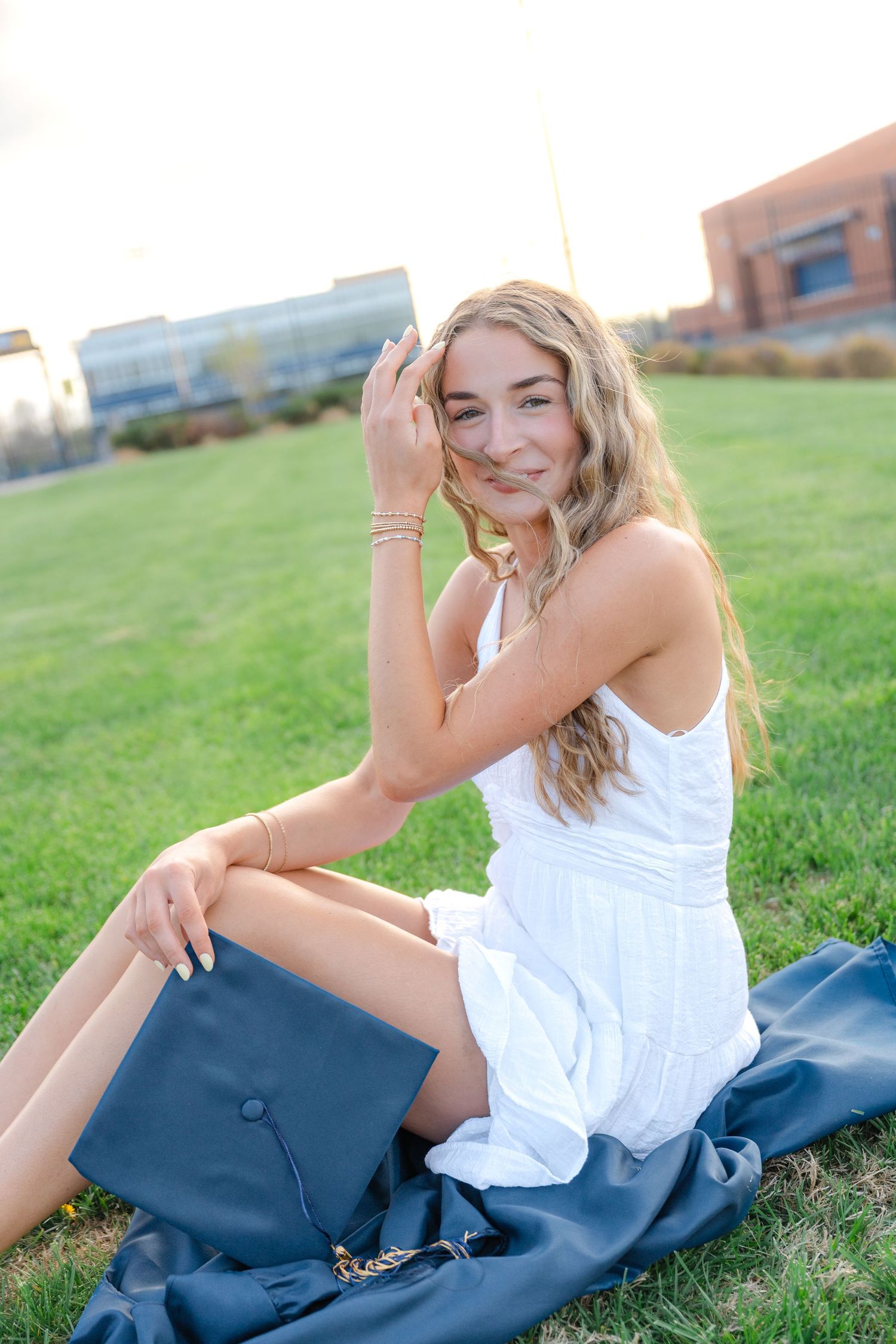 Young woman in white dress sitting on grass holding graduation cap, smiling at camera during golden hour.