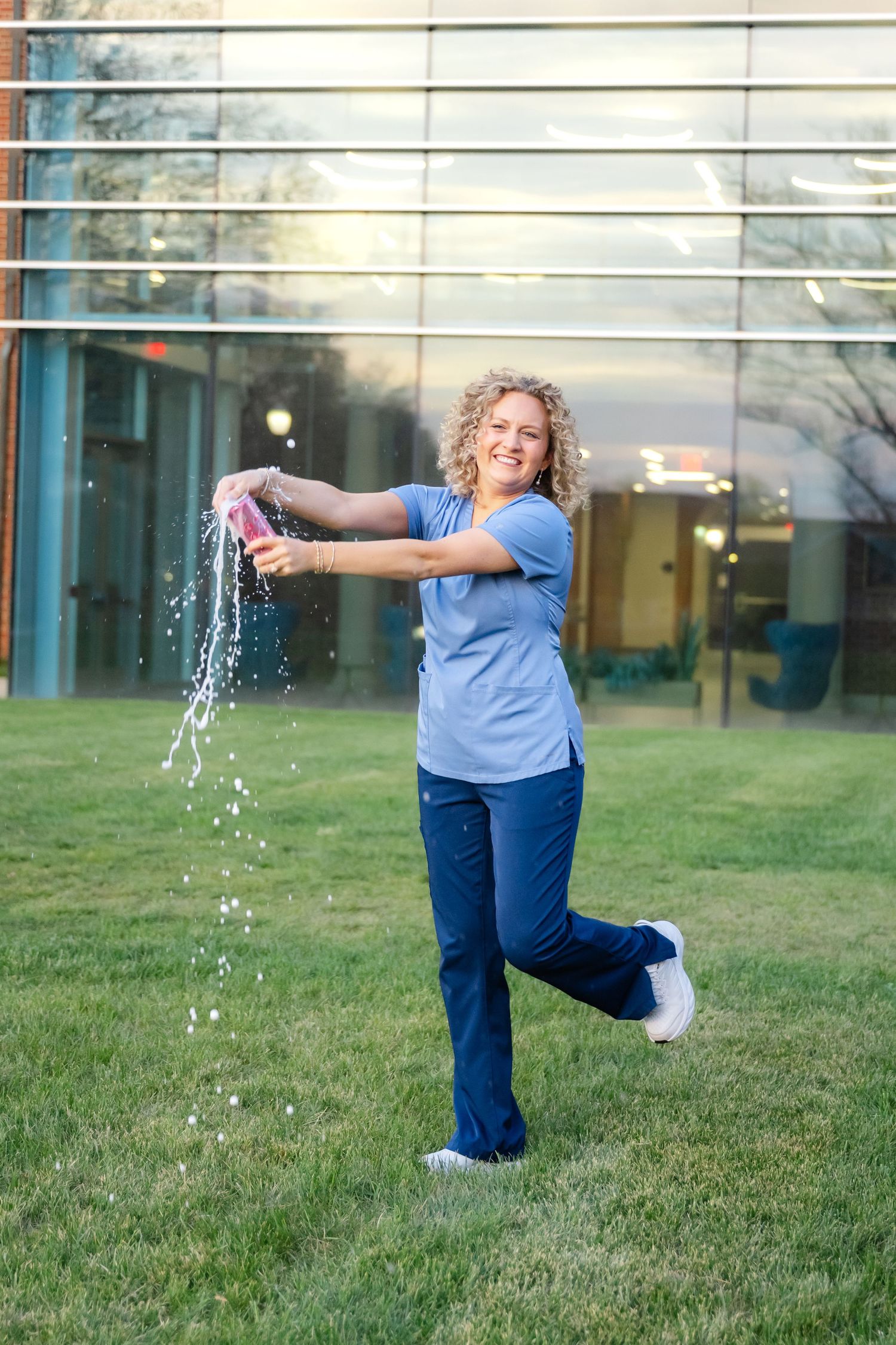 A smiling woman in blue scrubs playfully squirts water from a pink bottle on a green lawn outside a modern building.