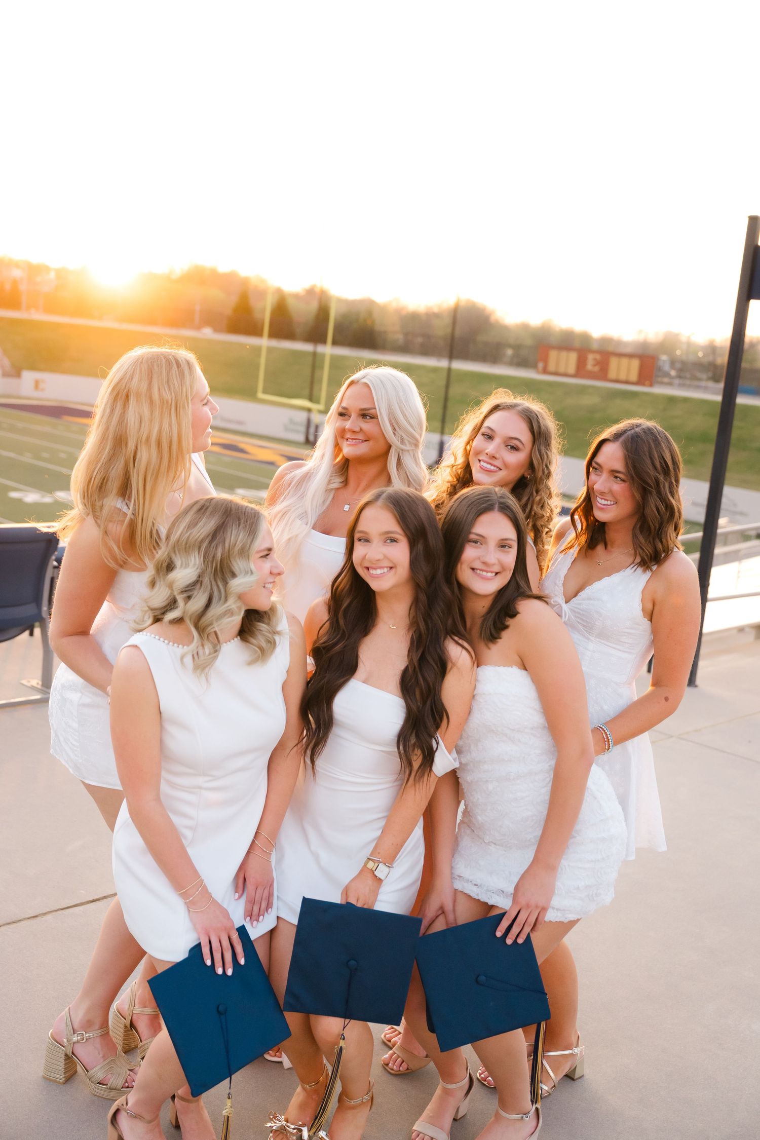 Seven young women in white dresses holding navy graduation caps pose together outdoors at sunset near a sports field.
