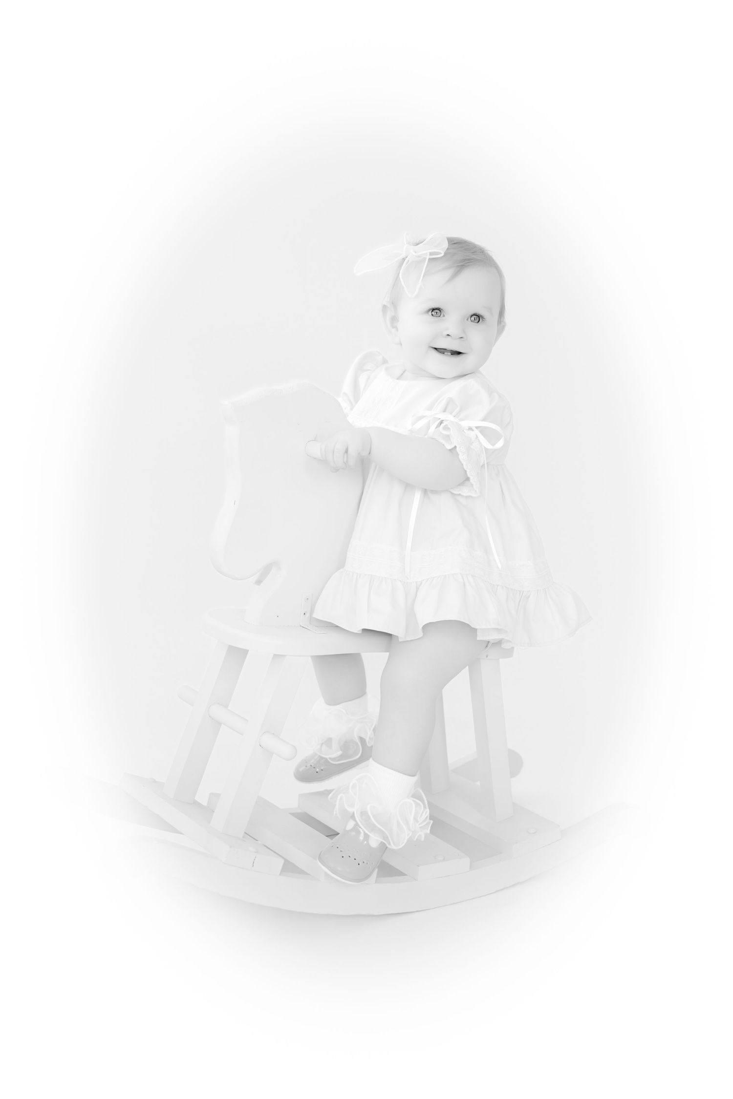 Black and white portrait of a smiling baby girl in white dress standing beside a wooden rocking horse.