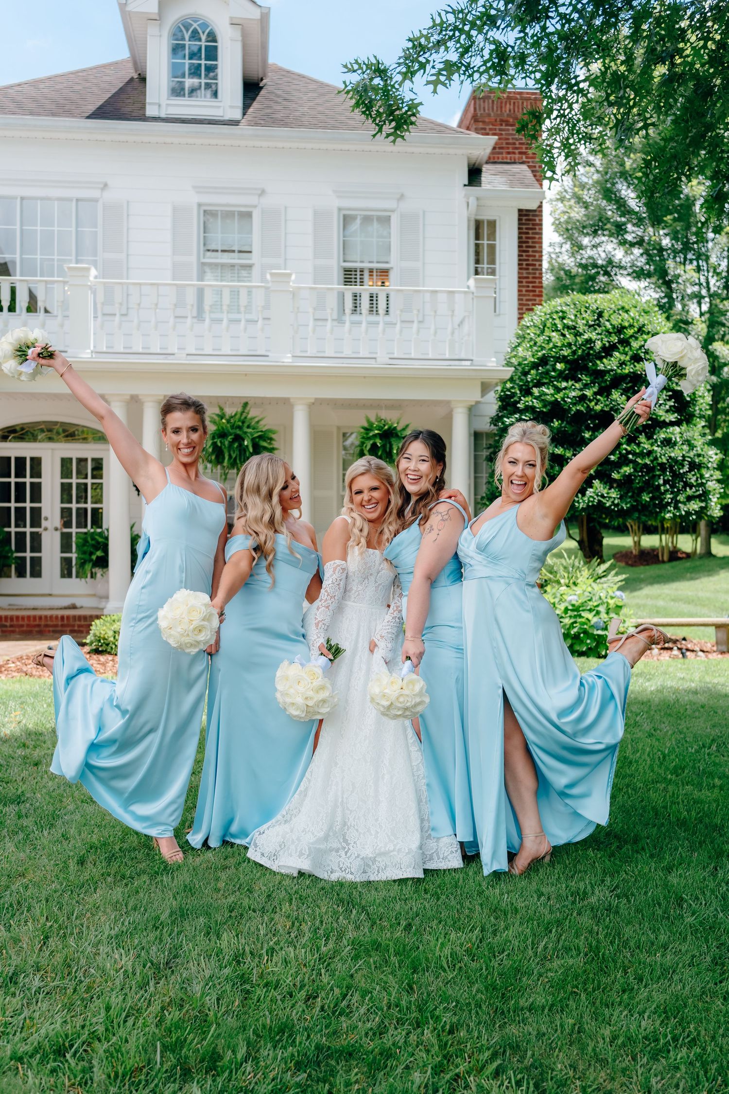 Wedding party in blue dresses and white gown poses joyfully on grass outside elegant white mansion.