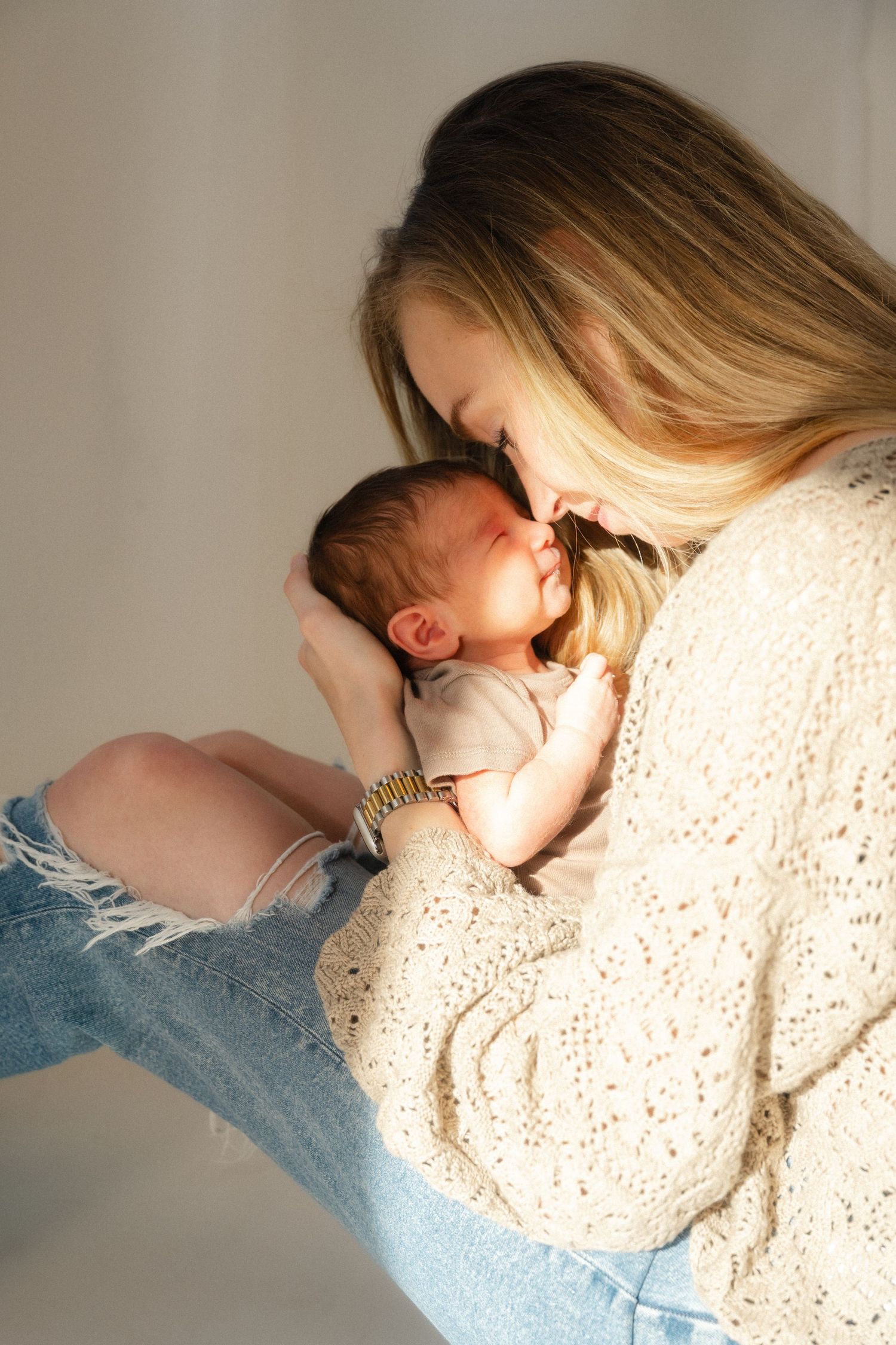 A young blonde mother in a cream sweater and ripped jeans tenderly holds her sleeping newborn baby close.