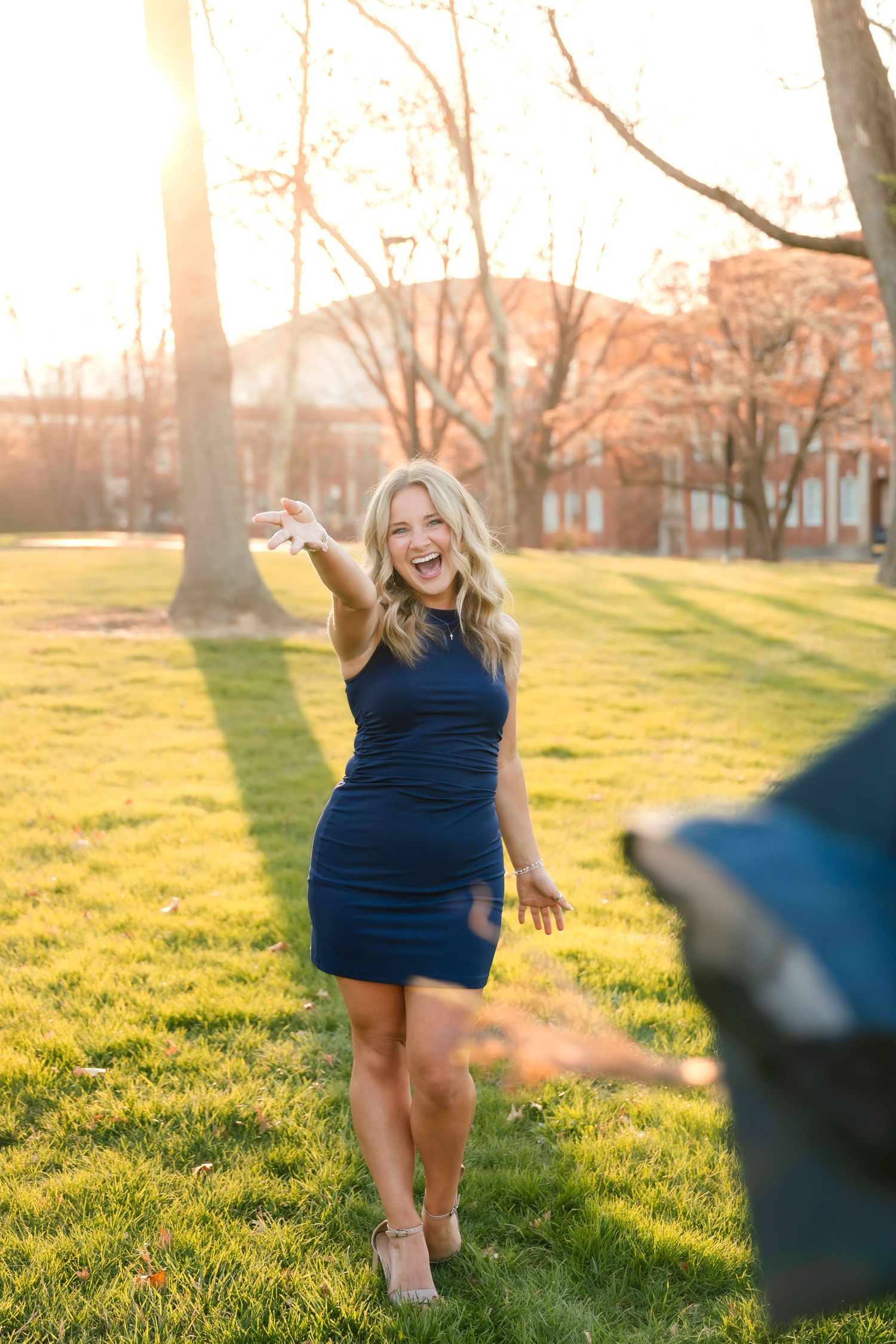 Laughing blonde woman in navy blue mini dress posing outdoors on green grass at golden hour sunset.