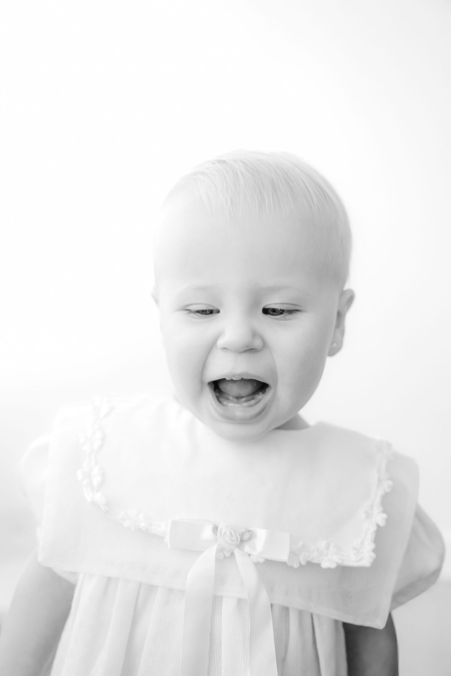 Black and white portrait of a happy laughing baby in a white dress with lace collar and bow.