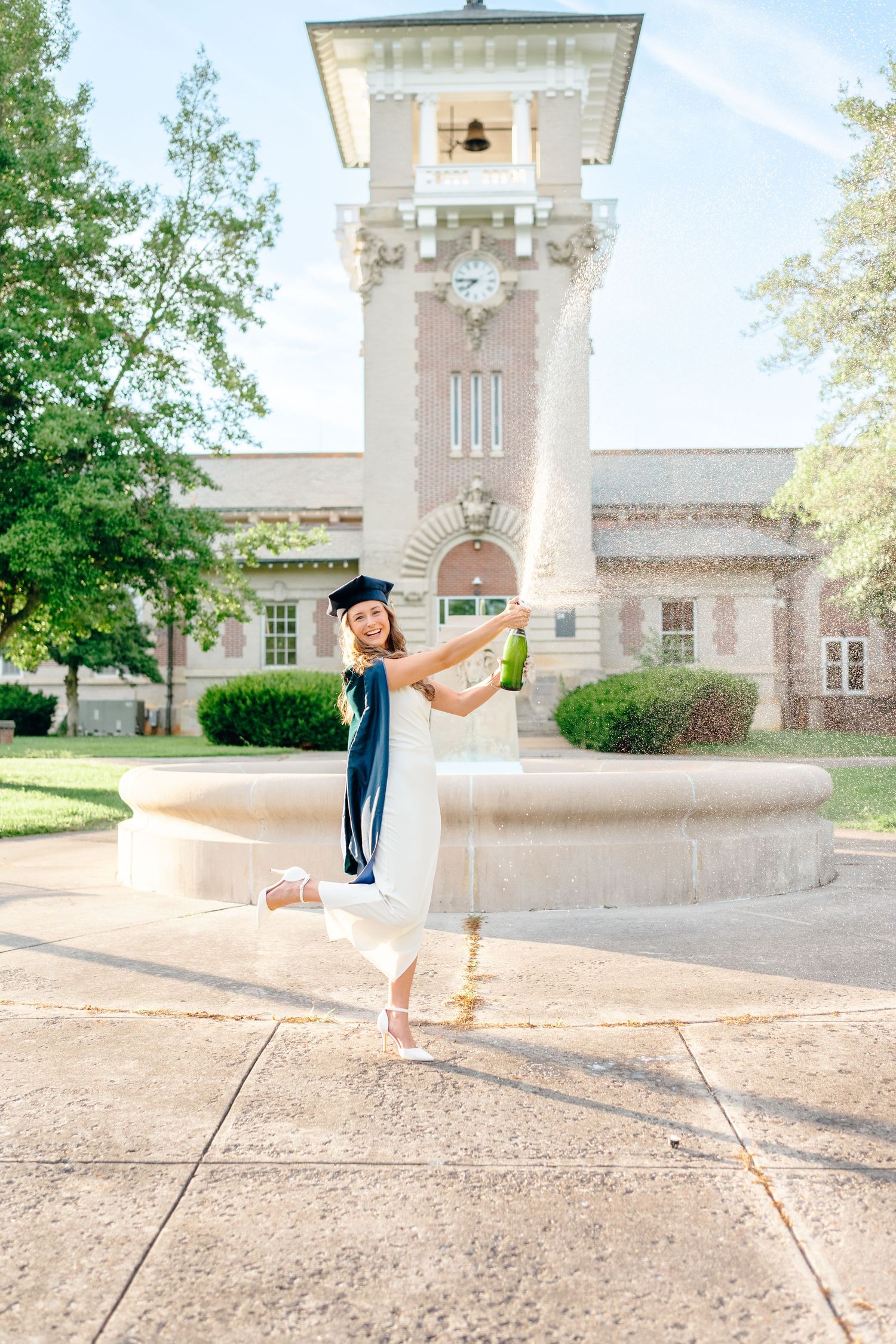 Graduate in blue cap and gown poses joyfully in front of a historic university building with clock tower.