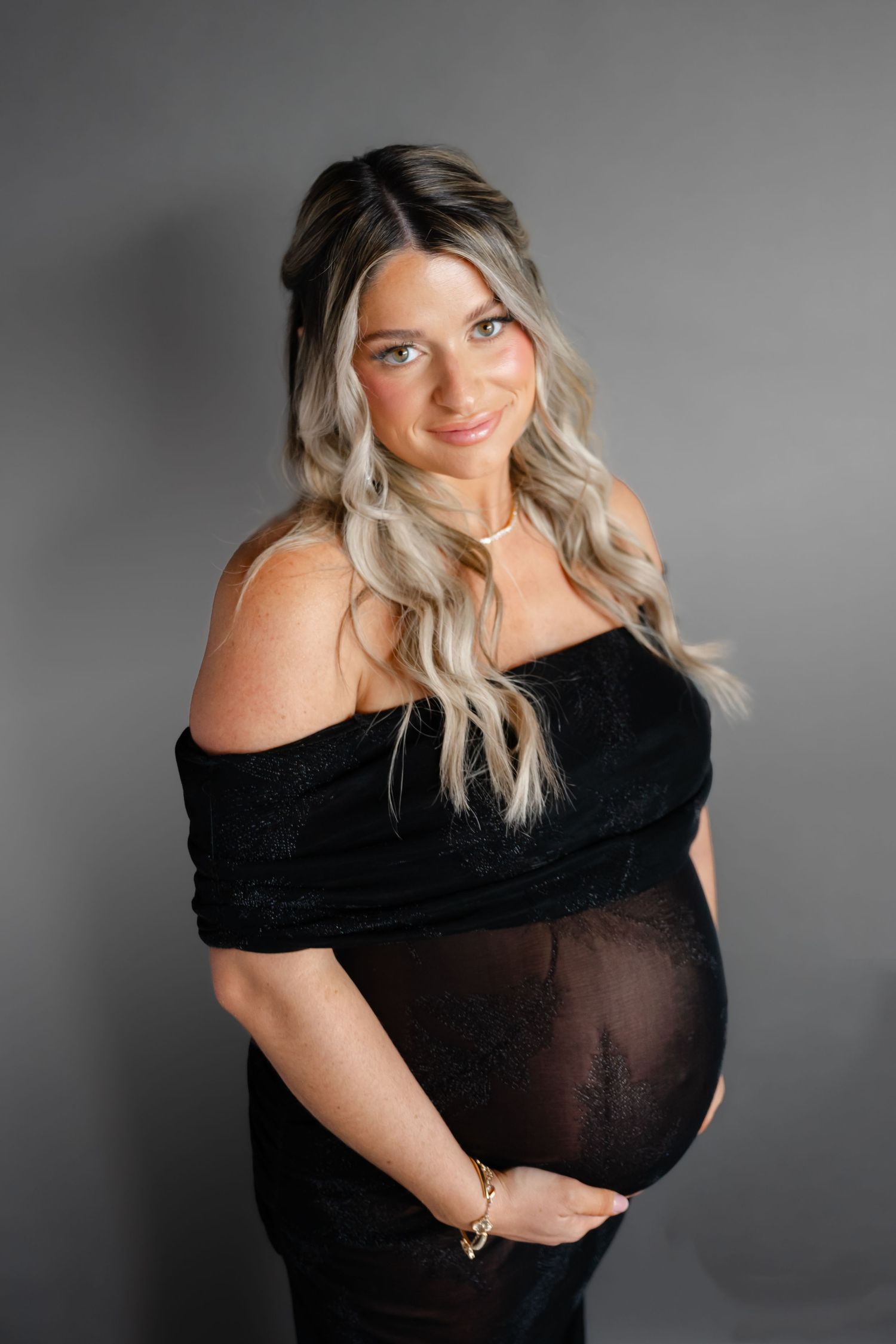 Maternity portrait featuring woman in black off-shoulder dress against grey background.
