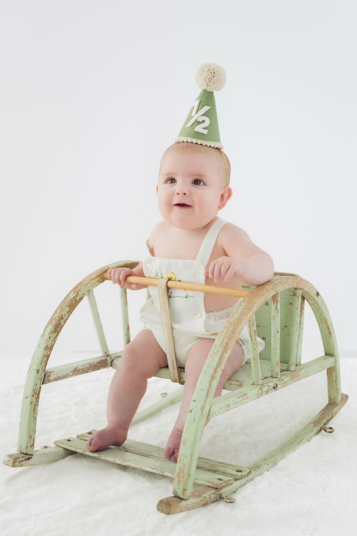Baby wearing party hat sits on vintage mint green sled against white background.