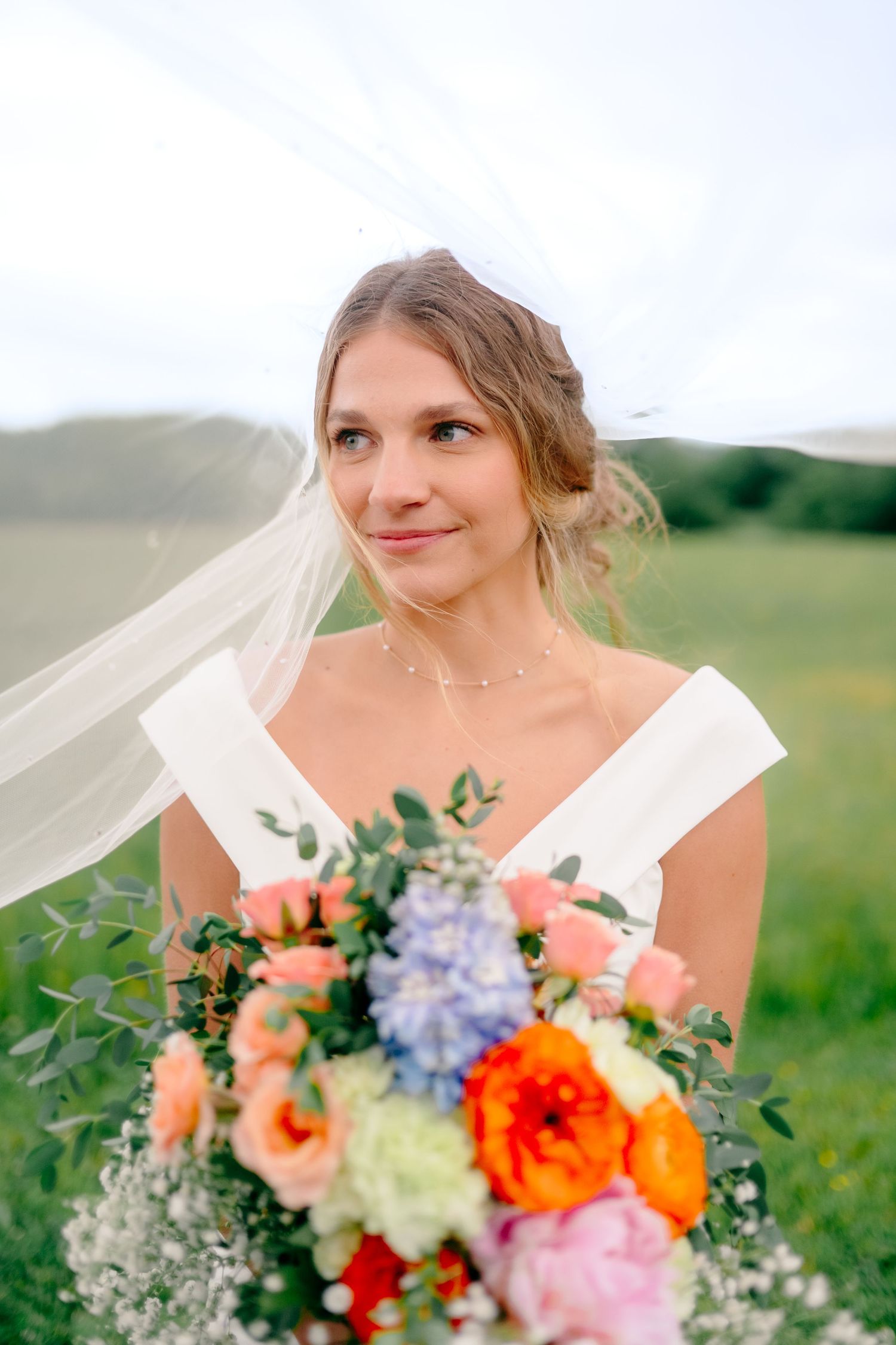 Bride with flowing veil holds vibrant bouquet of summer flowers in field.