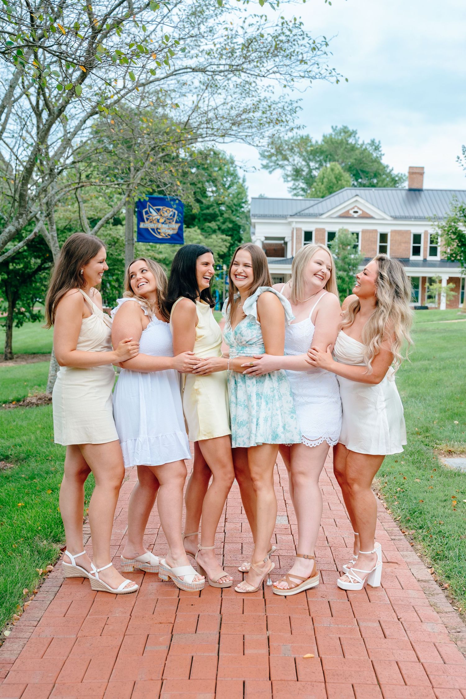 Group in white dresses pose together on brick pathway during graduation celebration.