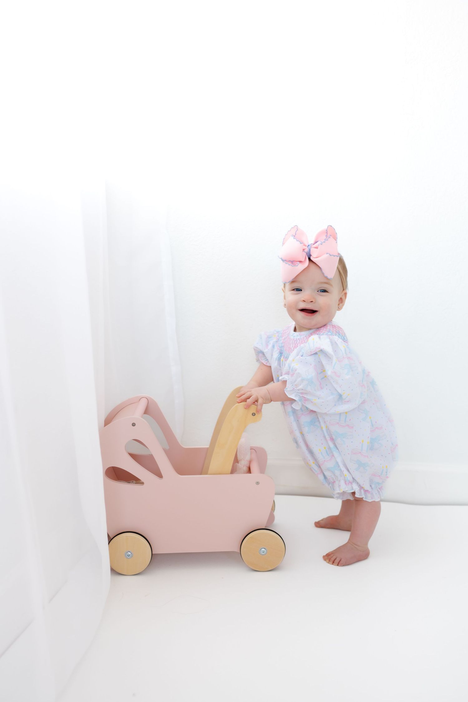 A happy baby in a floral dress plays with a pink wooden toy car on a white background.