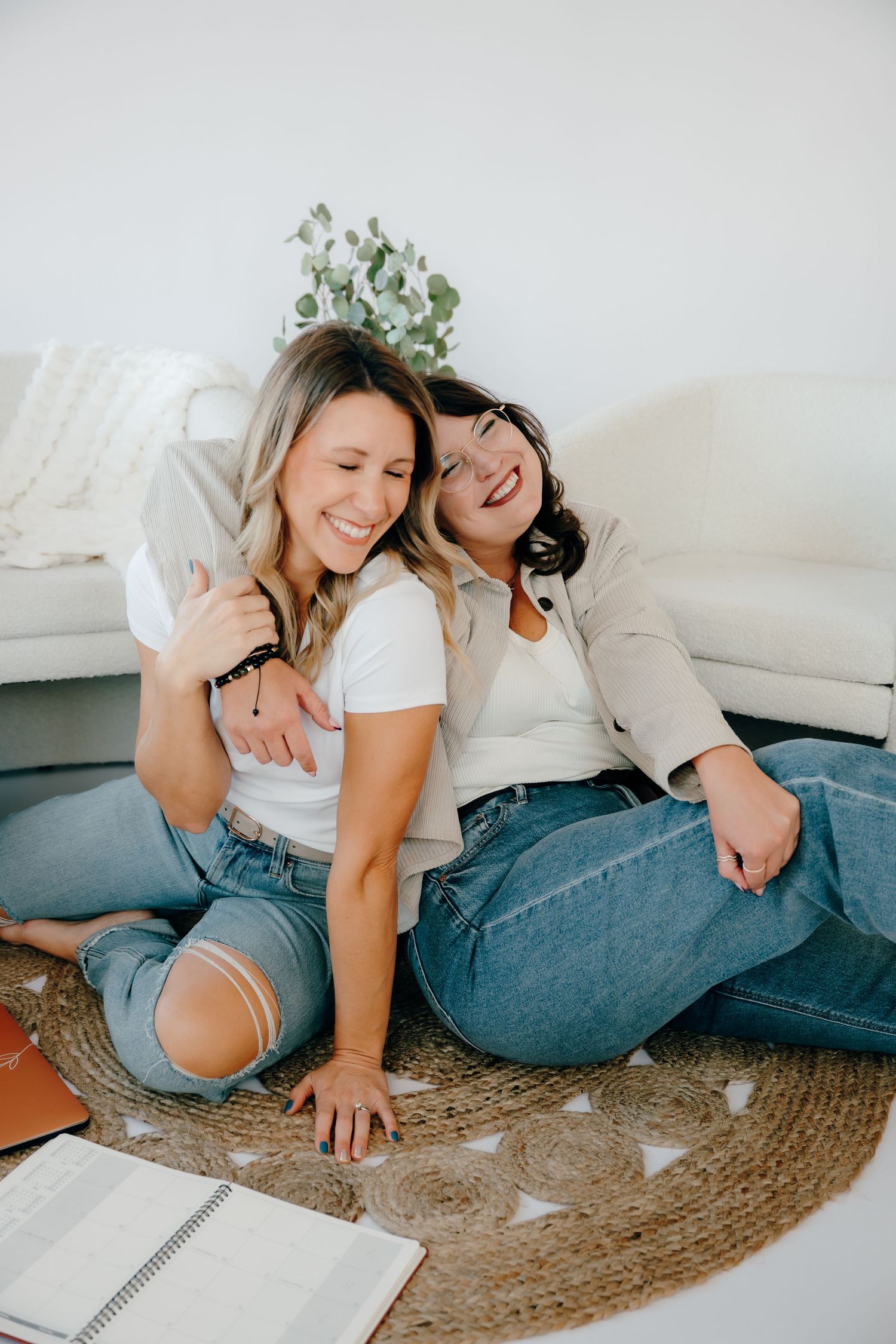 Two friends laughing and having fun while relaxing on a jute rug in casual denim outfits.