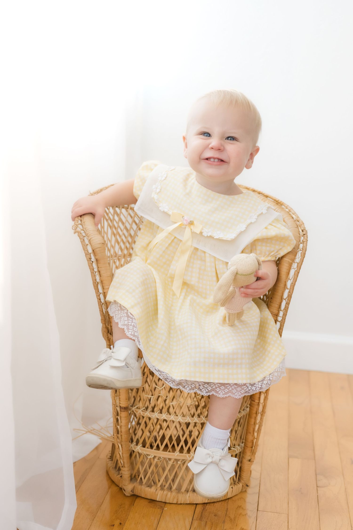 Smiling baby girl in yellow dress sitting in wicker chair holding stuffed toy, wearing white shoes and socks.