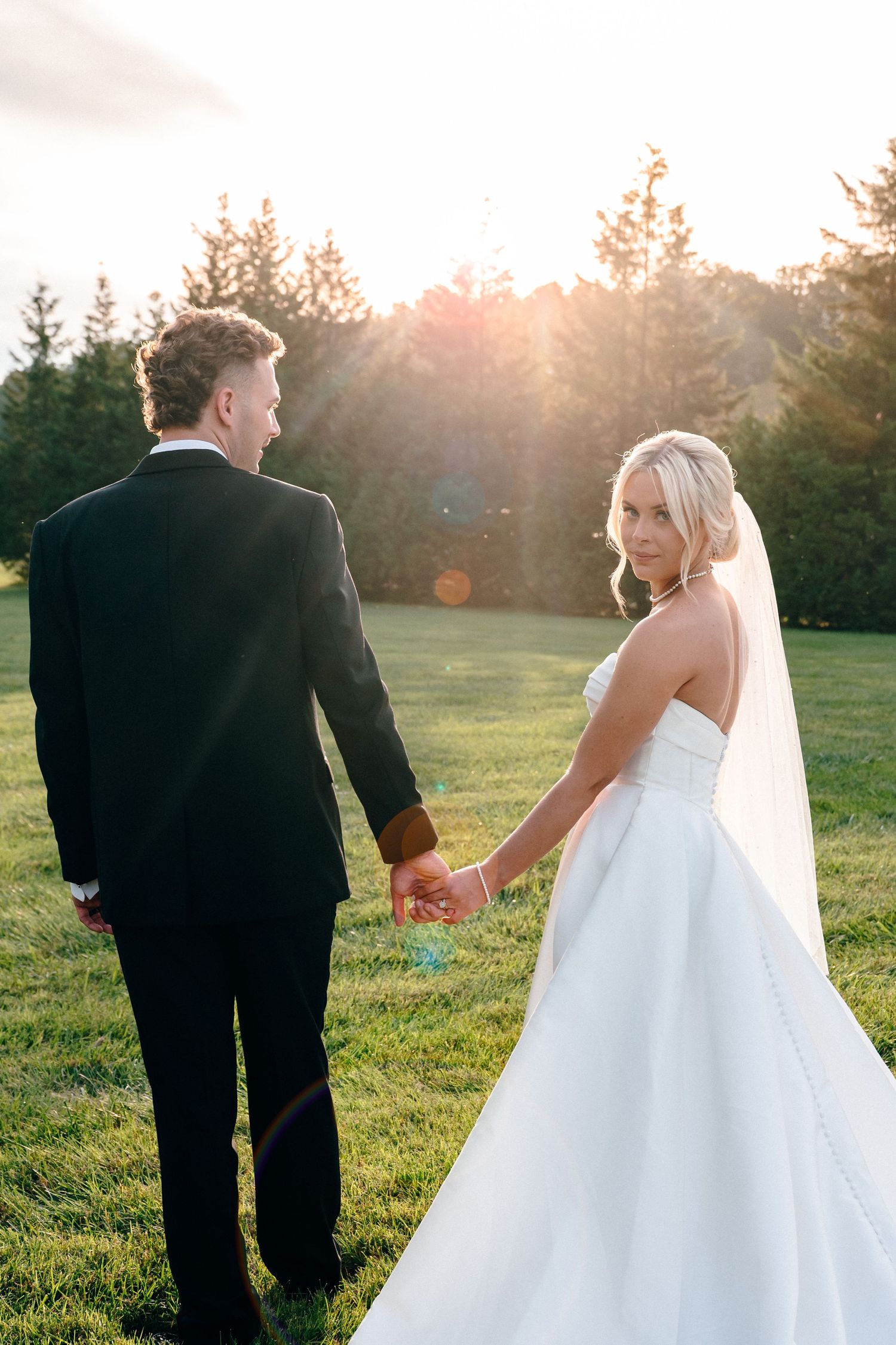 Newlyweds walk hand in hand through a sunlit field at sunset.
