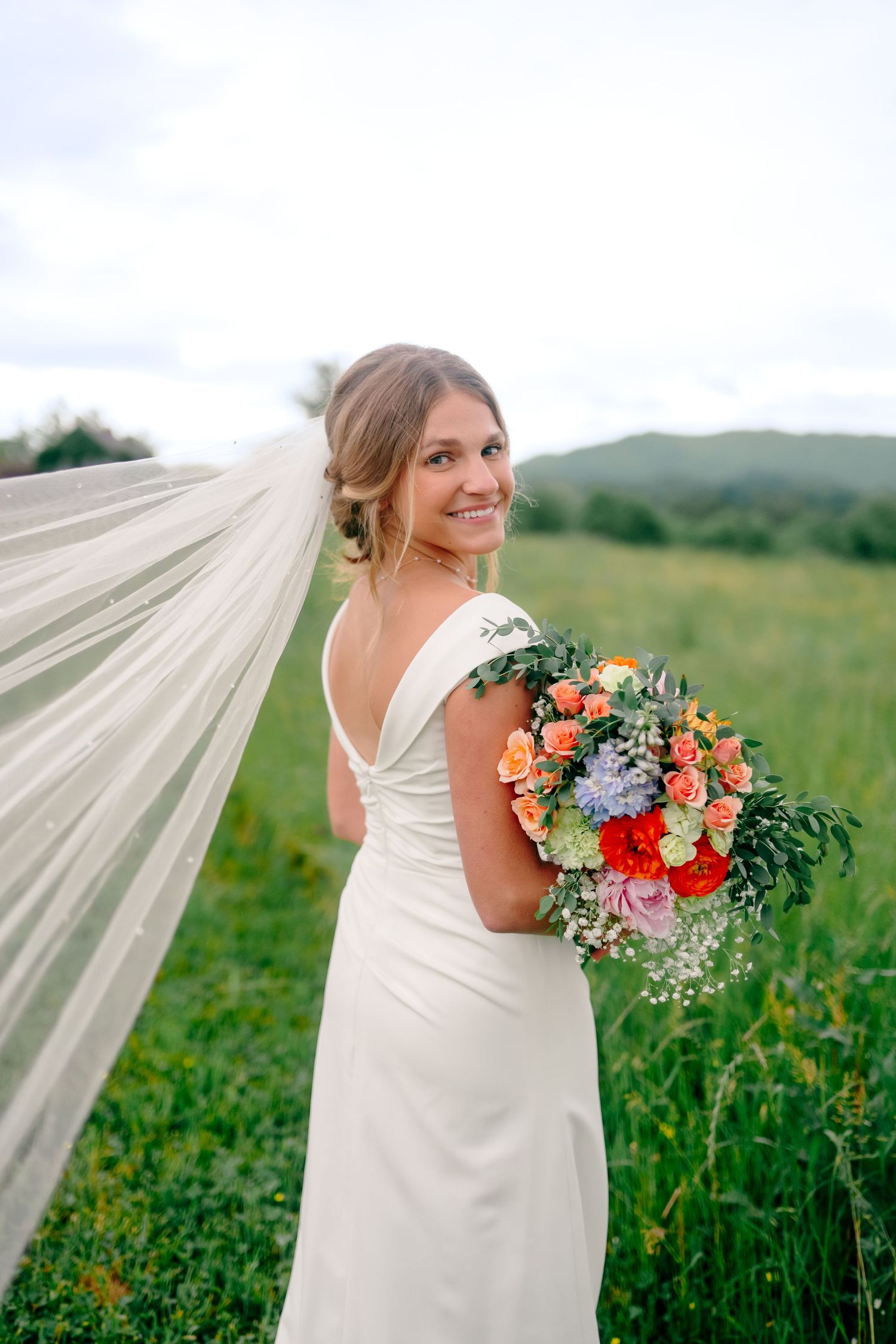 Bride with long veil holds colorful bouquet while smiling over shoulder in meadow.