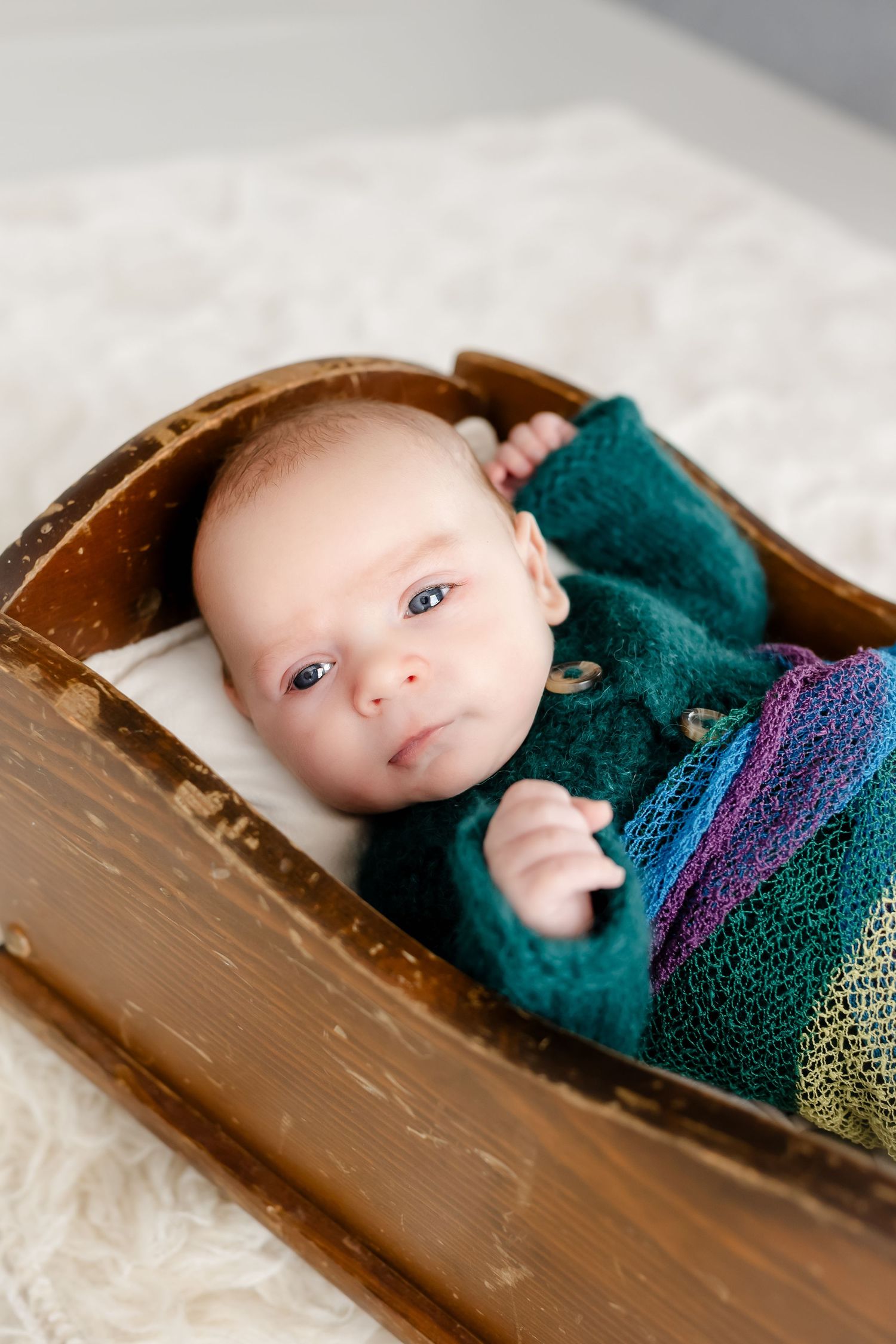 A small baby wearing a teal knit sweater lies in a rustic wooden drawer on white bedding.