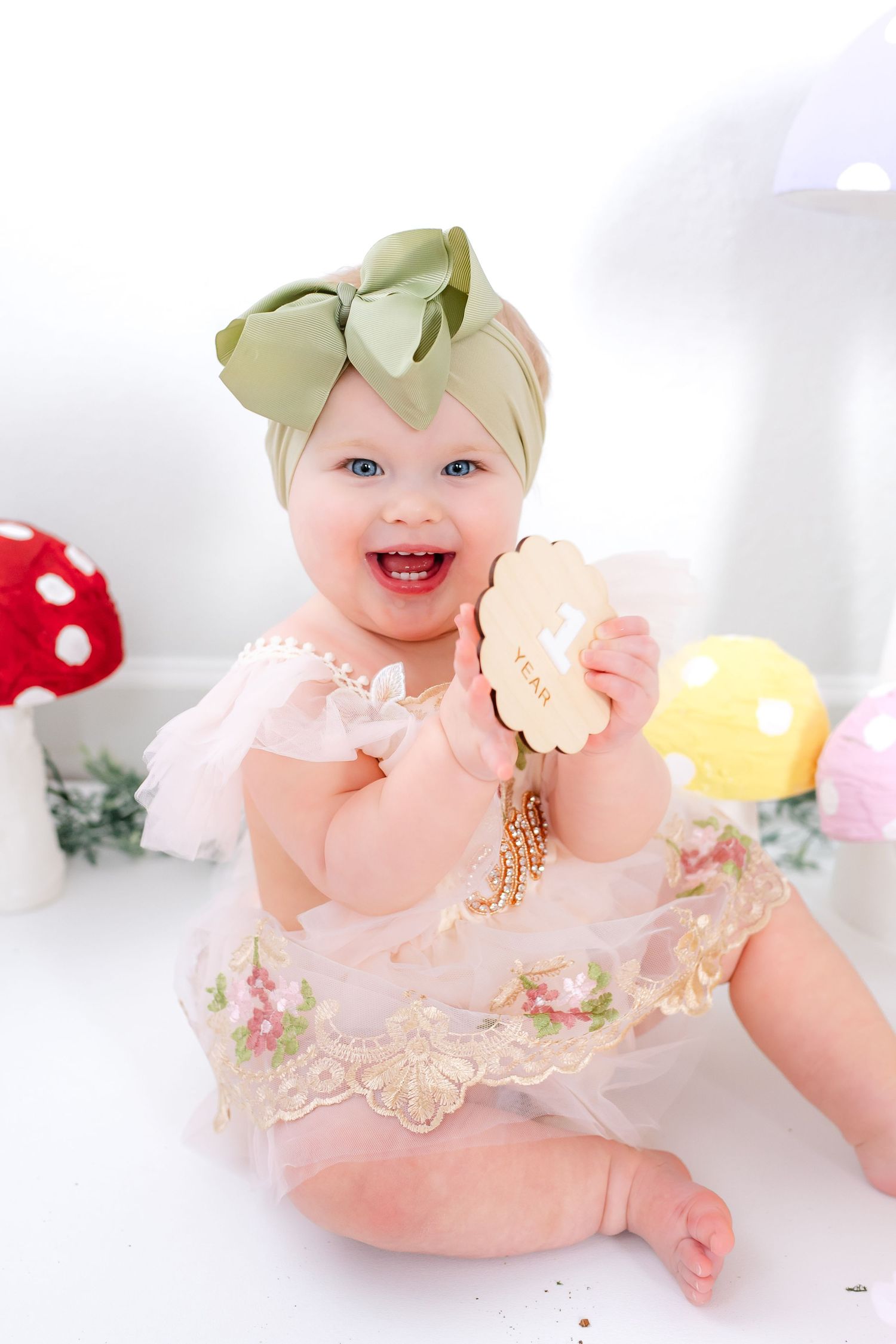 A baby wearing a peach dress and green bow headband sits on a white background with floral decorations.