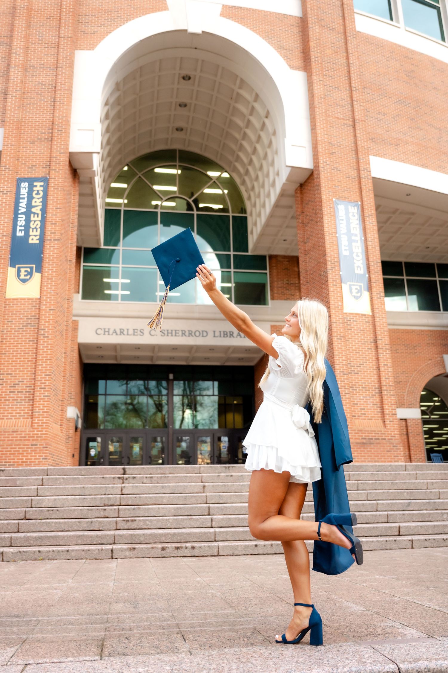 Blonde graduate in white dress tosses cap outside Charles Sherrod Library, holding navy gown, celebrating graduation day.