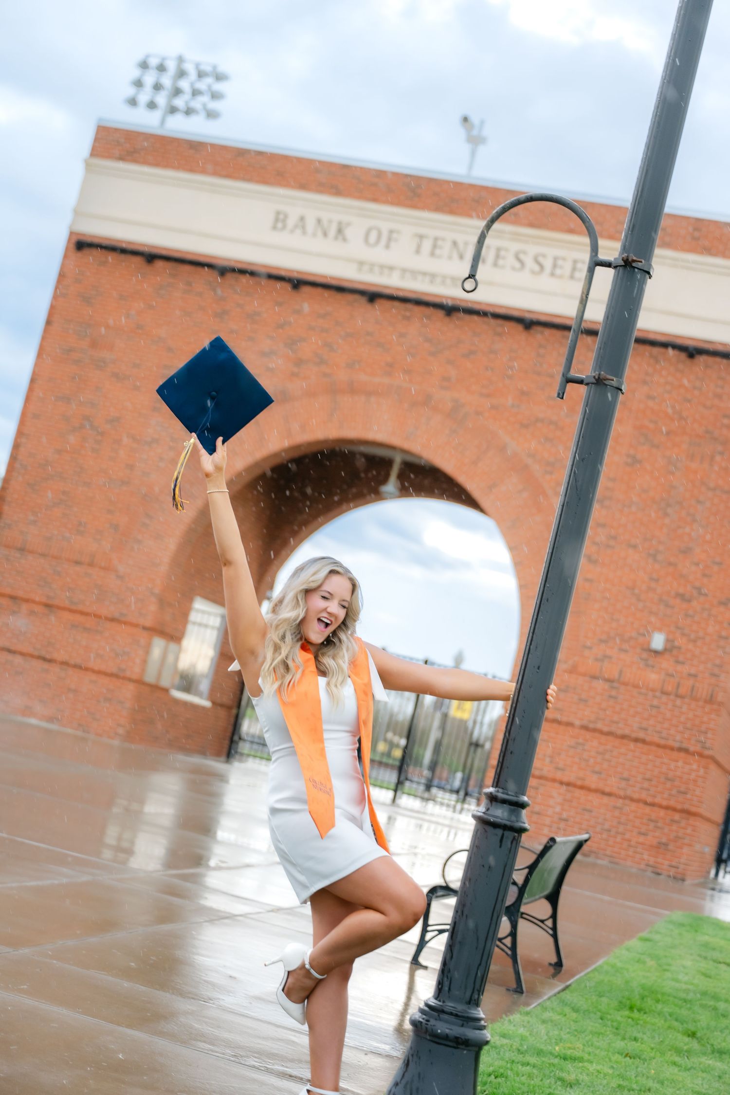 Excited female graduate in white dress and orange sash raises cap outside Bank of Tennessee stadium on rainy day.