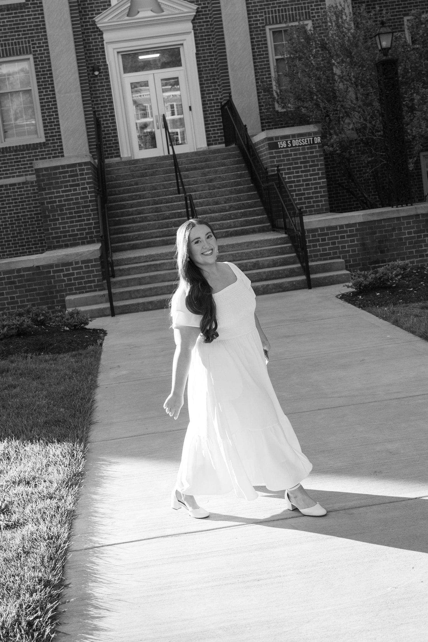 Black and white photo of a smiling woman in a white dress walking on a sunny sidewalk outside a brick building.