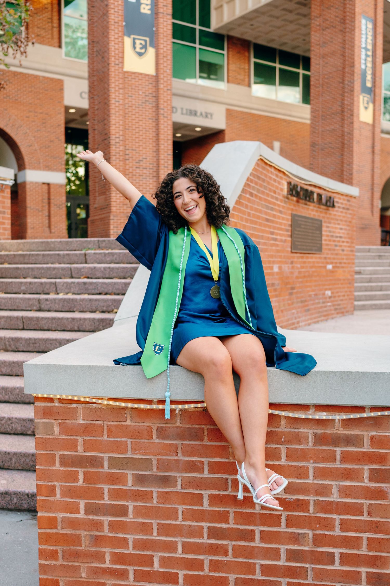 Graduate in blue dress and green stole poses on brick steps of university building.