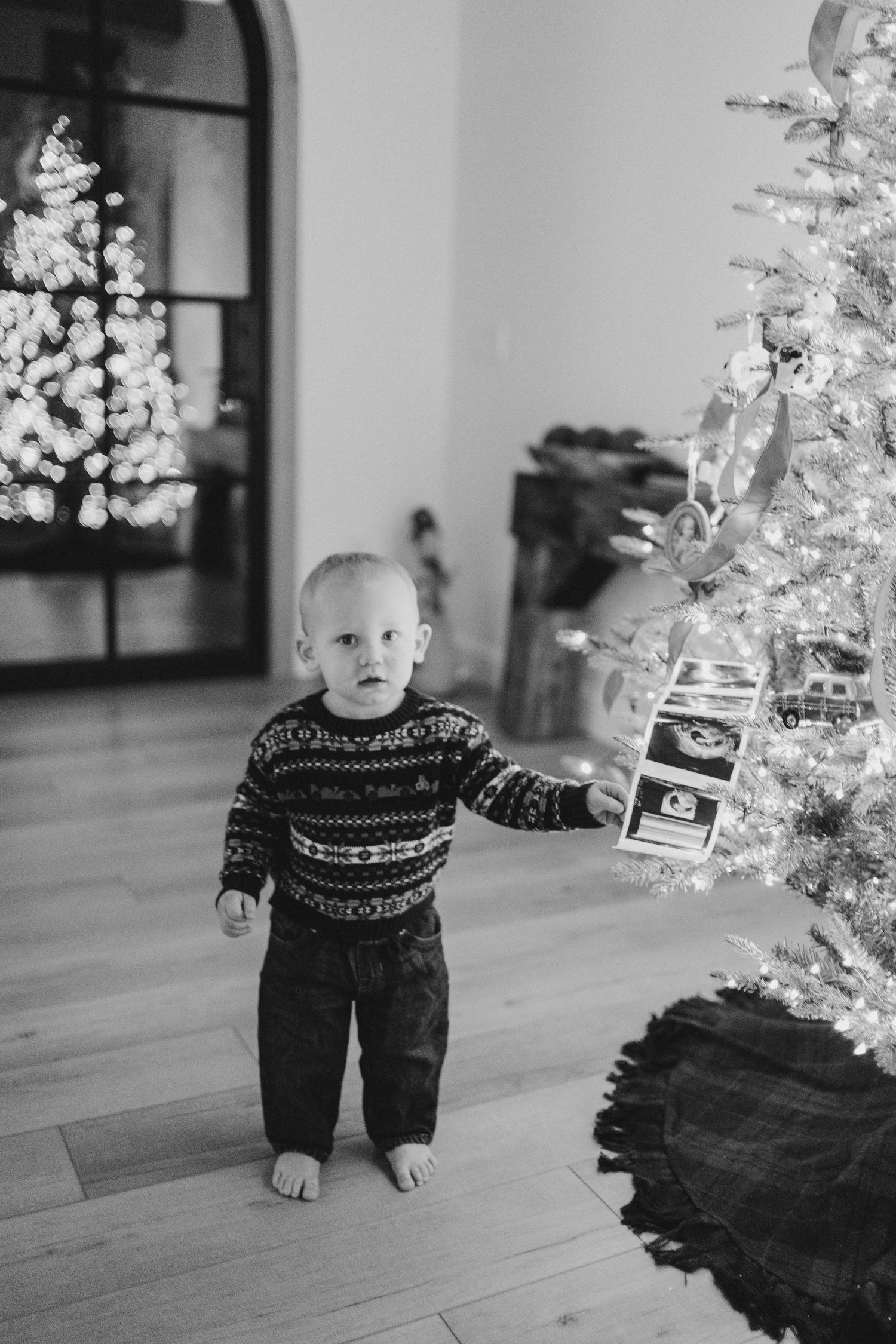 Black and white photos of baby in patterned sweater playing near illuminated Christmas trees.