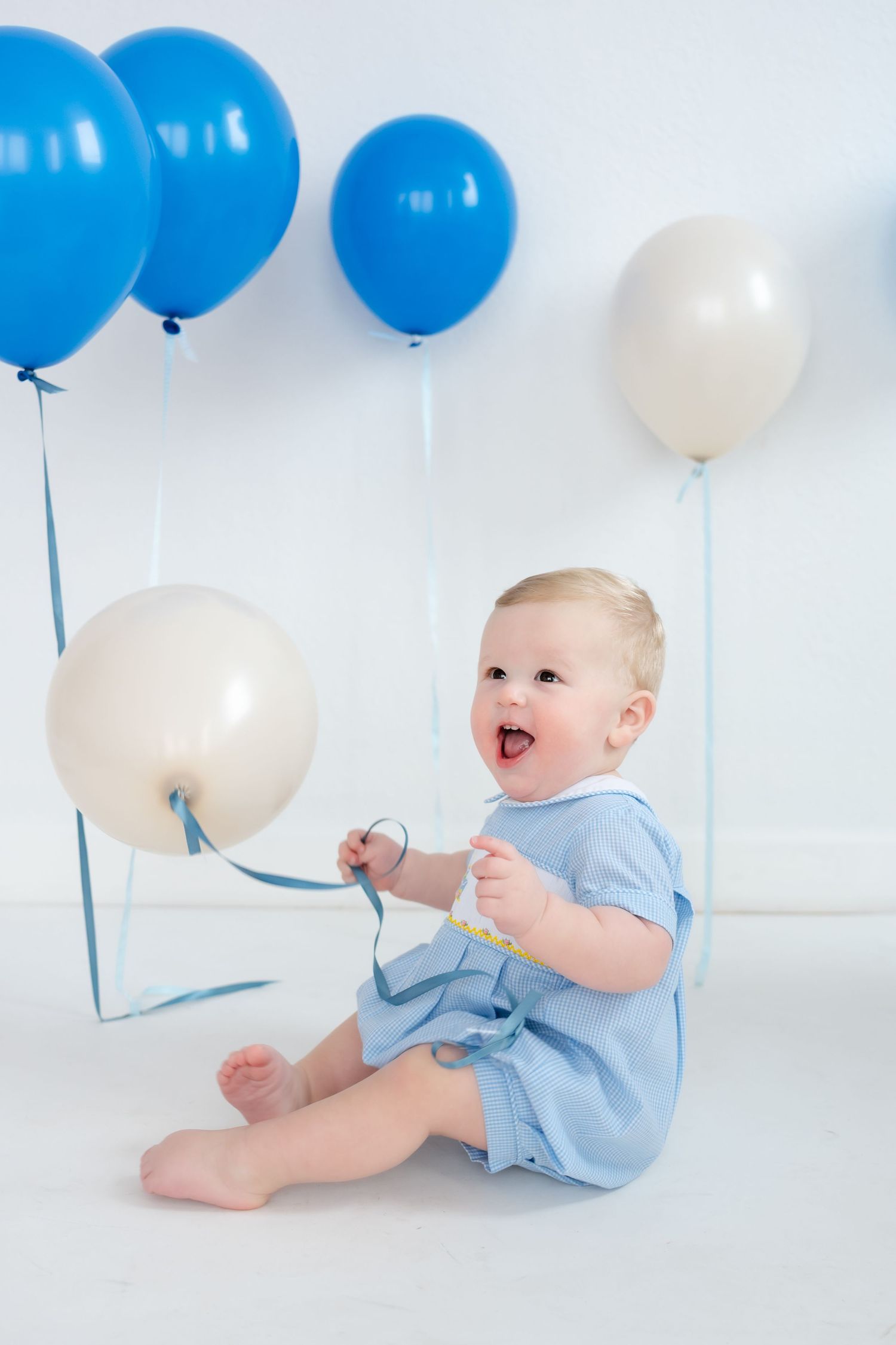 Baby in light blue outfit sits on white floor with blue and white balloons during birthday or celebration photoshoot.