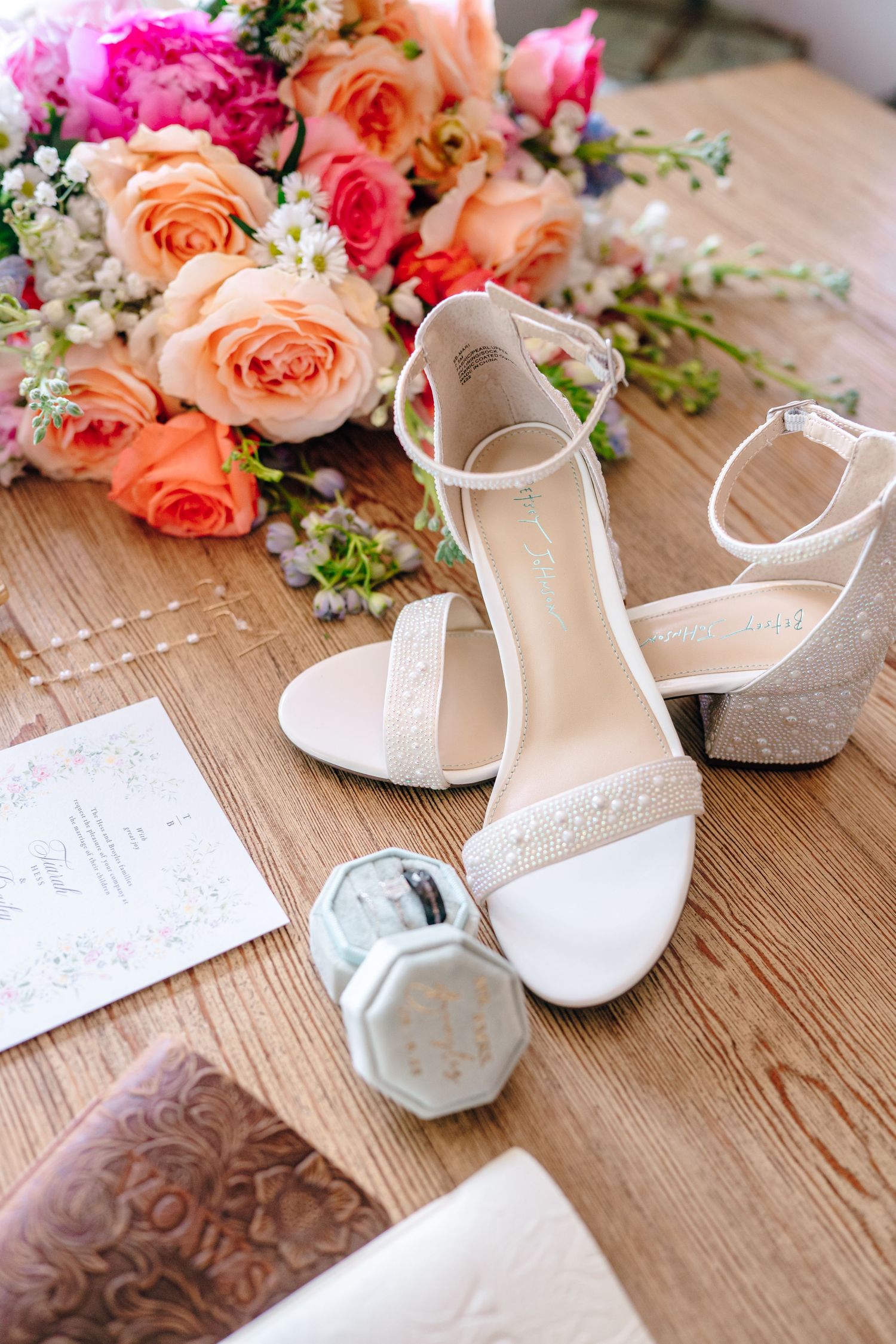 Elegant white bridal sandals with coral and pink roses and wedding stationery on wooden surface.