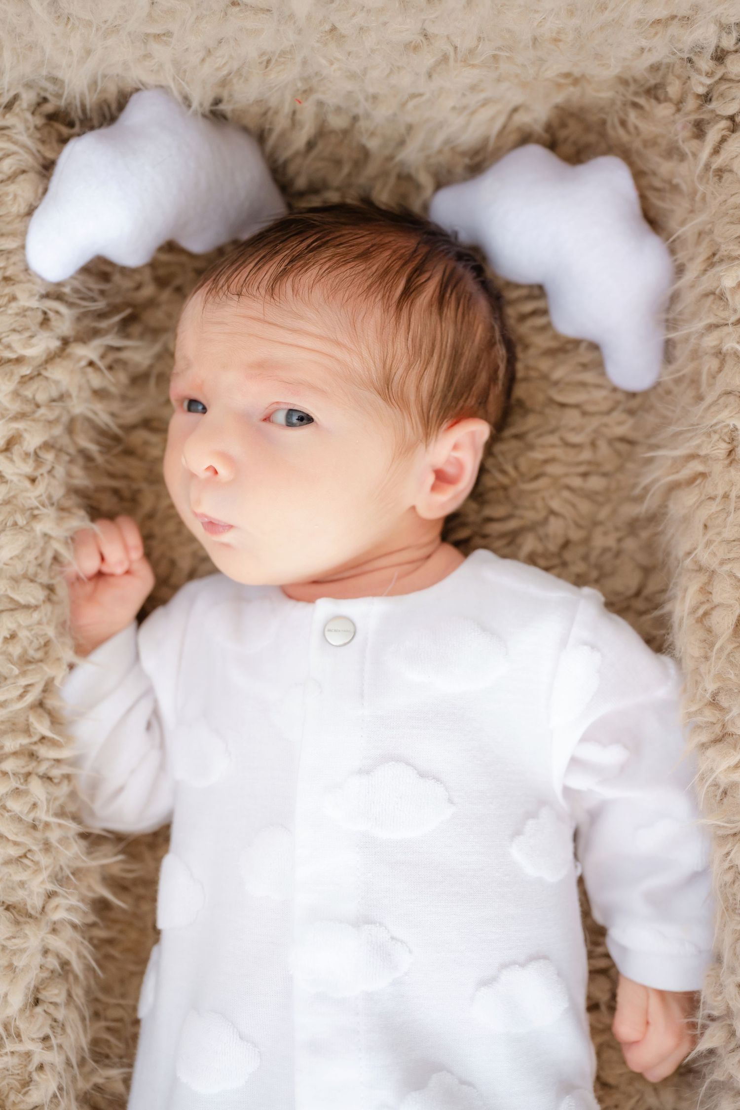 Newborn baby in white cloud-print onesie lying on fluffy beige blanket with star-shaped pillows.