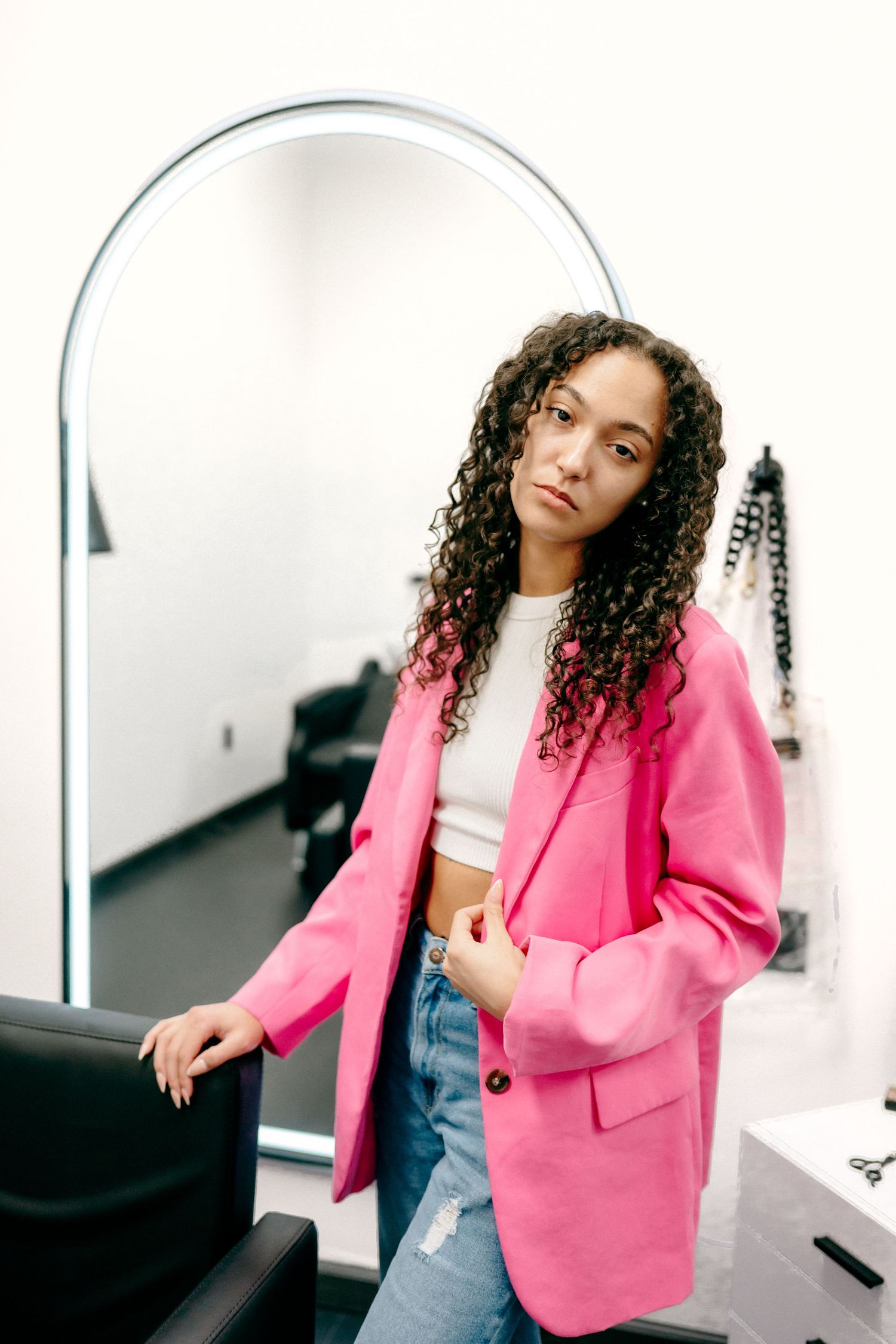 Person wearing pink blazer and crop top leans against salon chair near arched mirror.