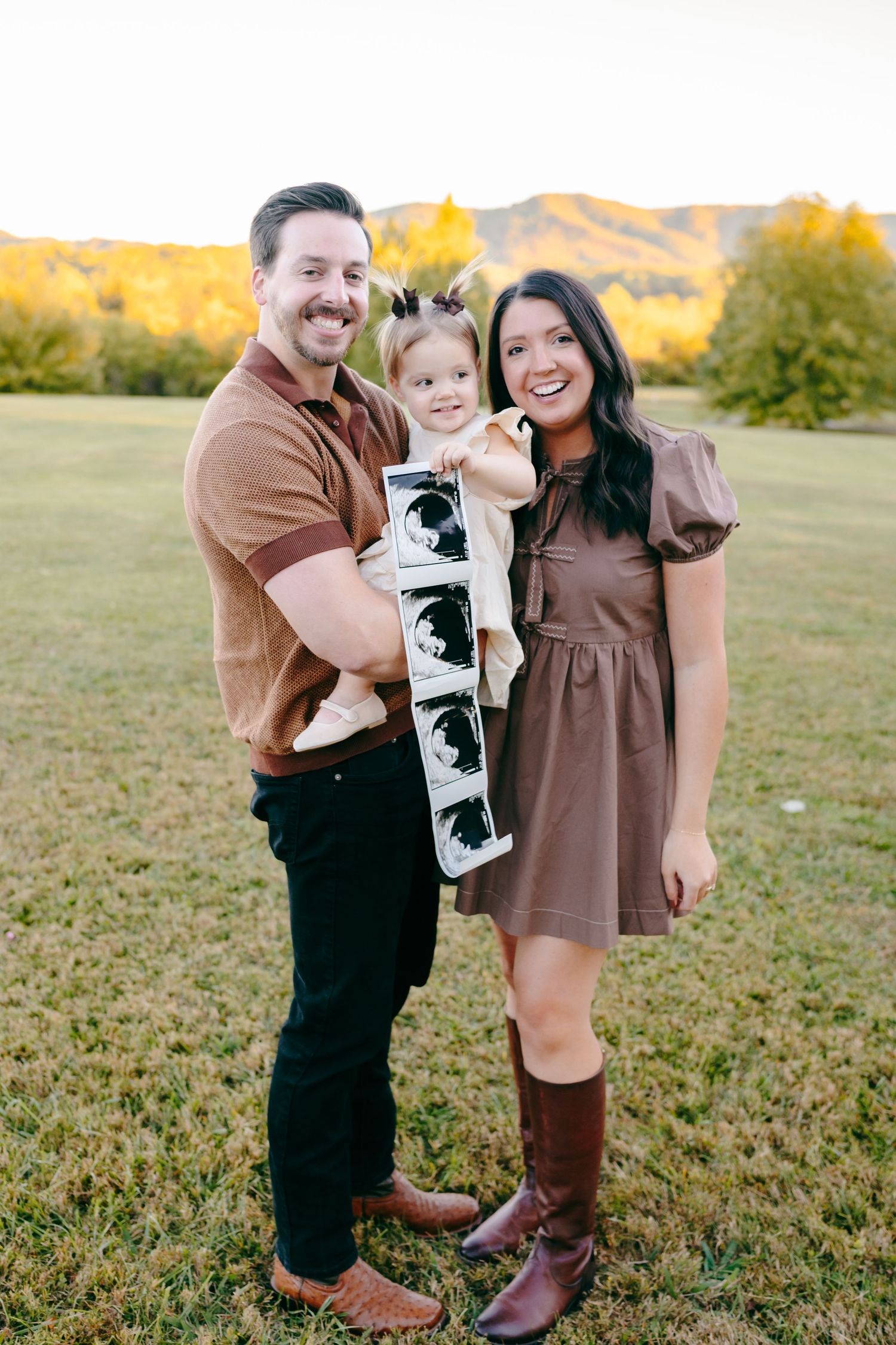 A family poses together in a grassy field during golden hour wearing brown and black outfits.