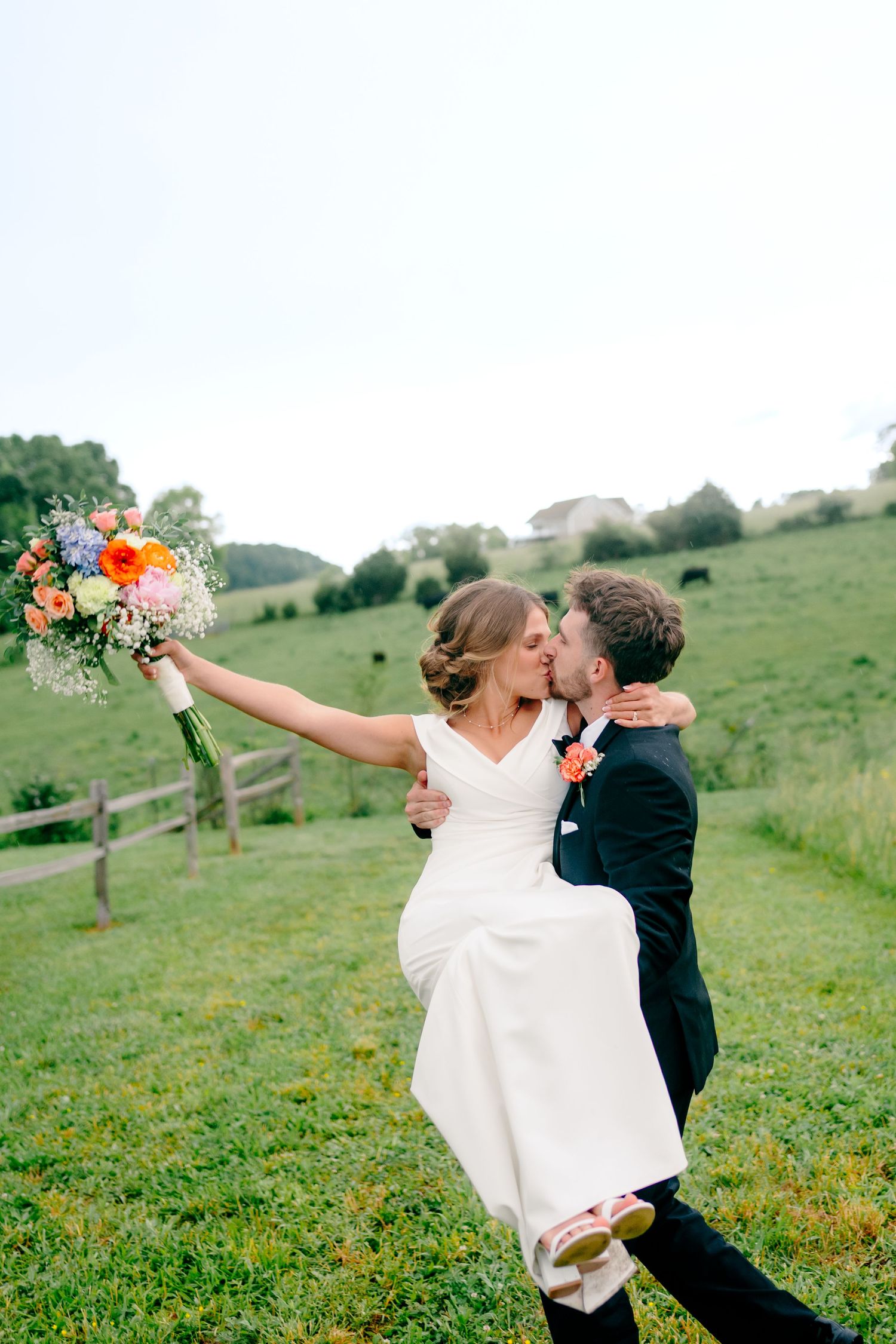 Newlywed couple embraces in a romantic outdoor photoshoot on a green hillside with colorful bouquet.