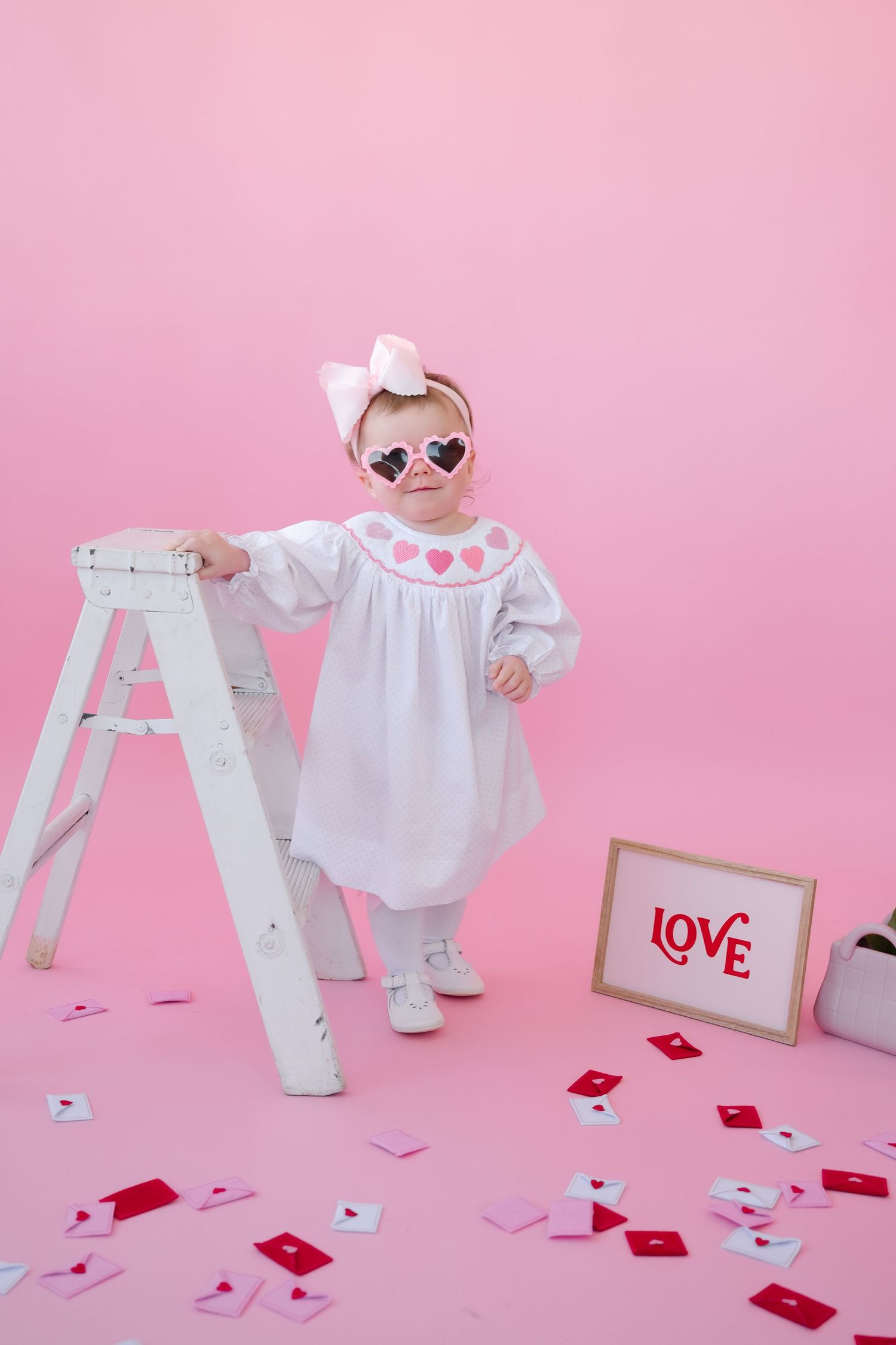 Small child in white dress and sunglasses stands by ladder with Valentine's Day decor on pink background.