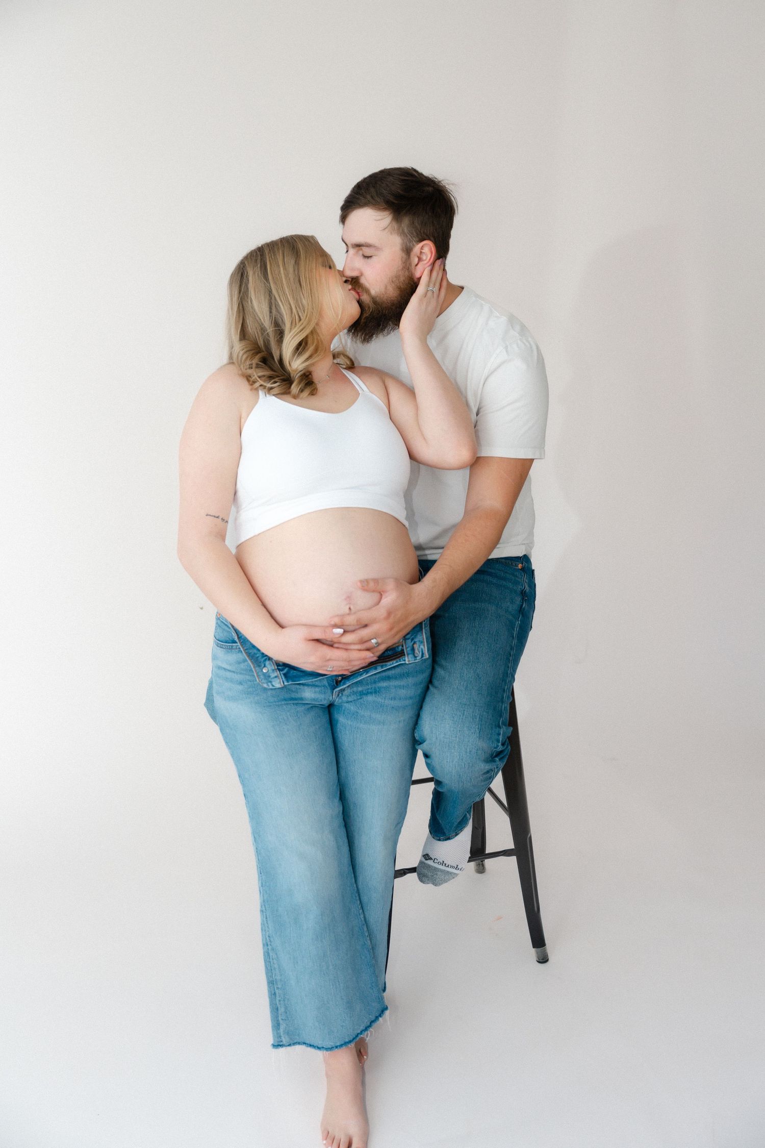 Intimate maternity photo of a couple in casual attire sharing a tender moment against a white backdrop.