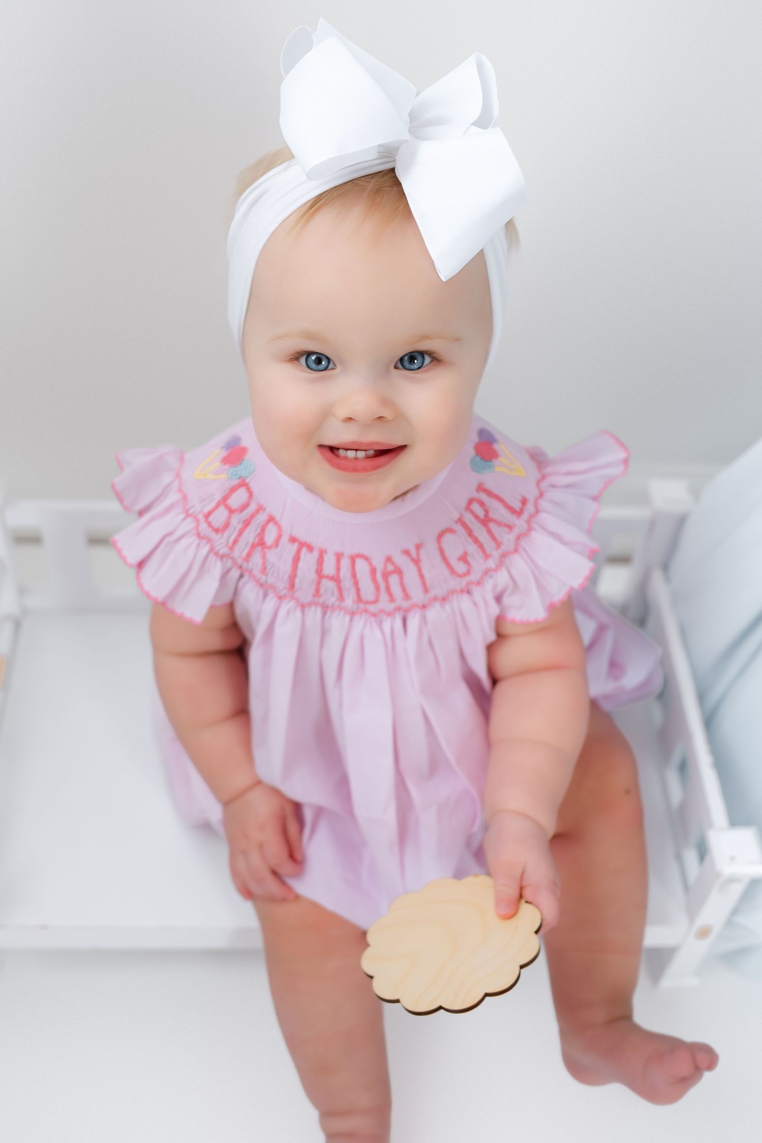 Baby in pink ruffled dress and white bow headband smiles while sitting in bright white setting.