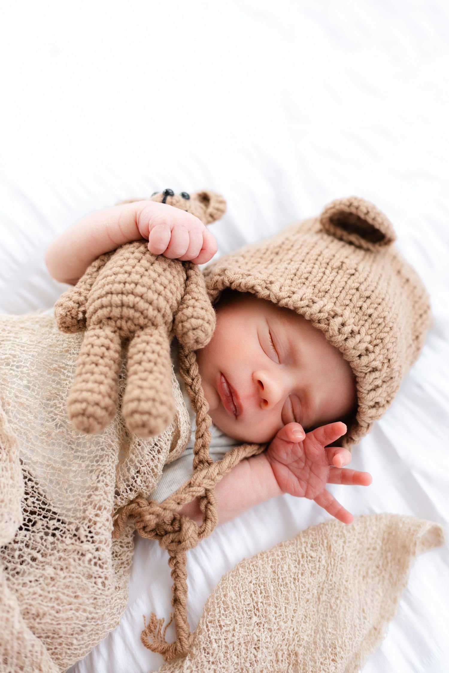 Sleeping newborn baby wearing cozy beige knit bear hat and wrap on white bedding.