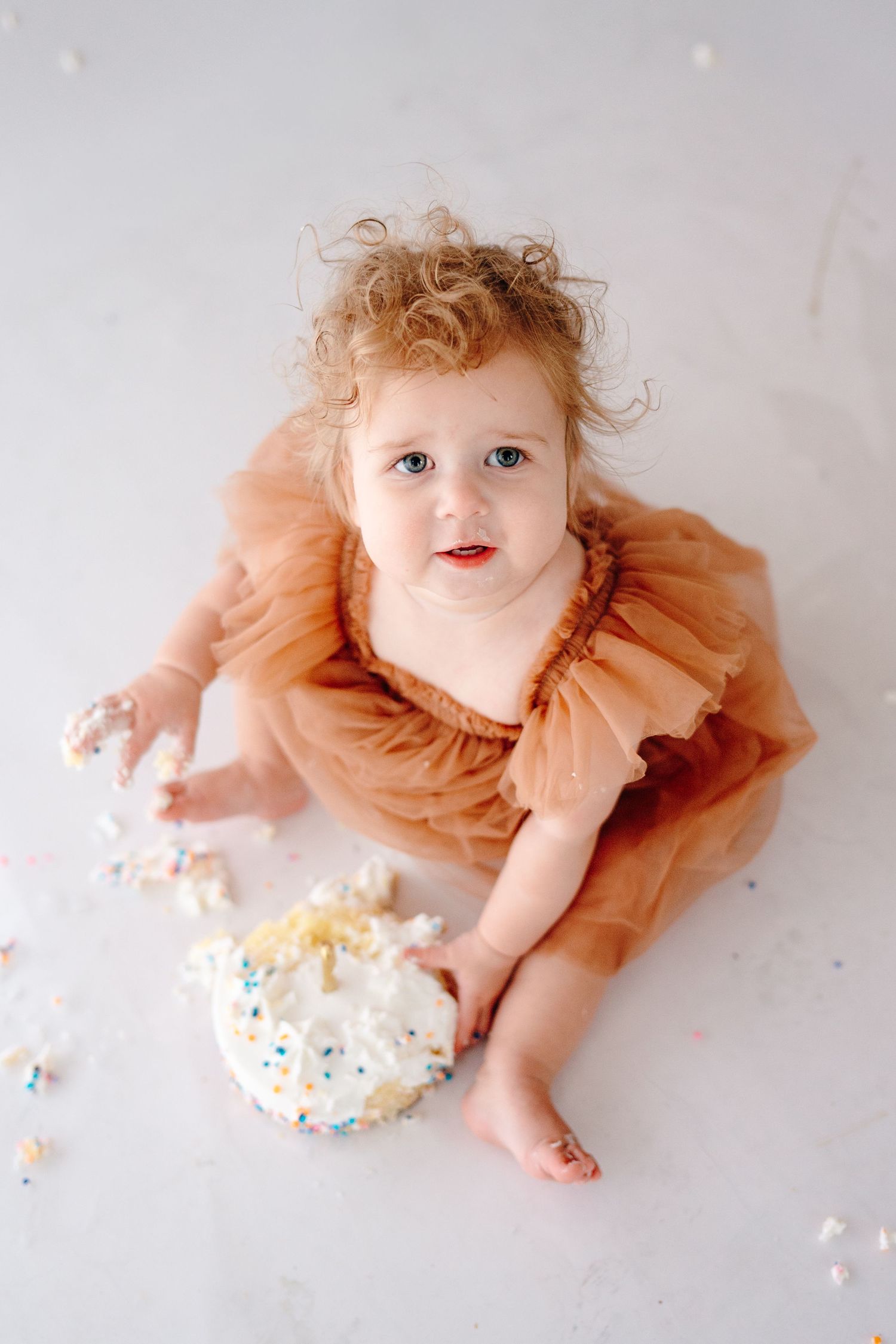 Baby in peach tulle dress explores white frosted cake with sprinkles.