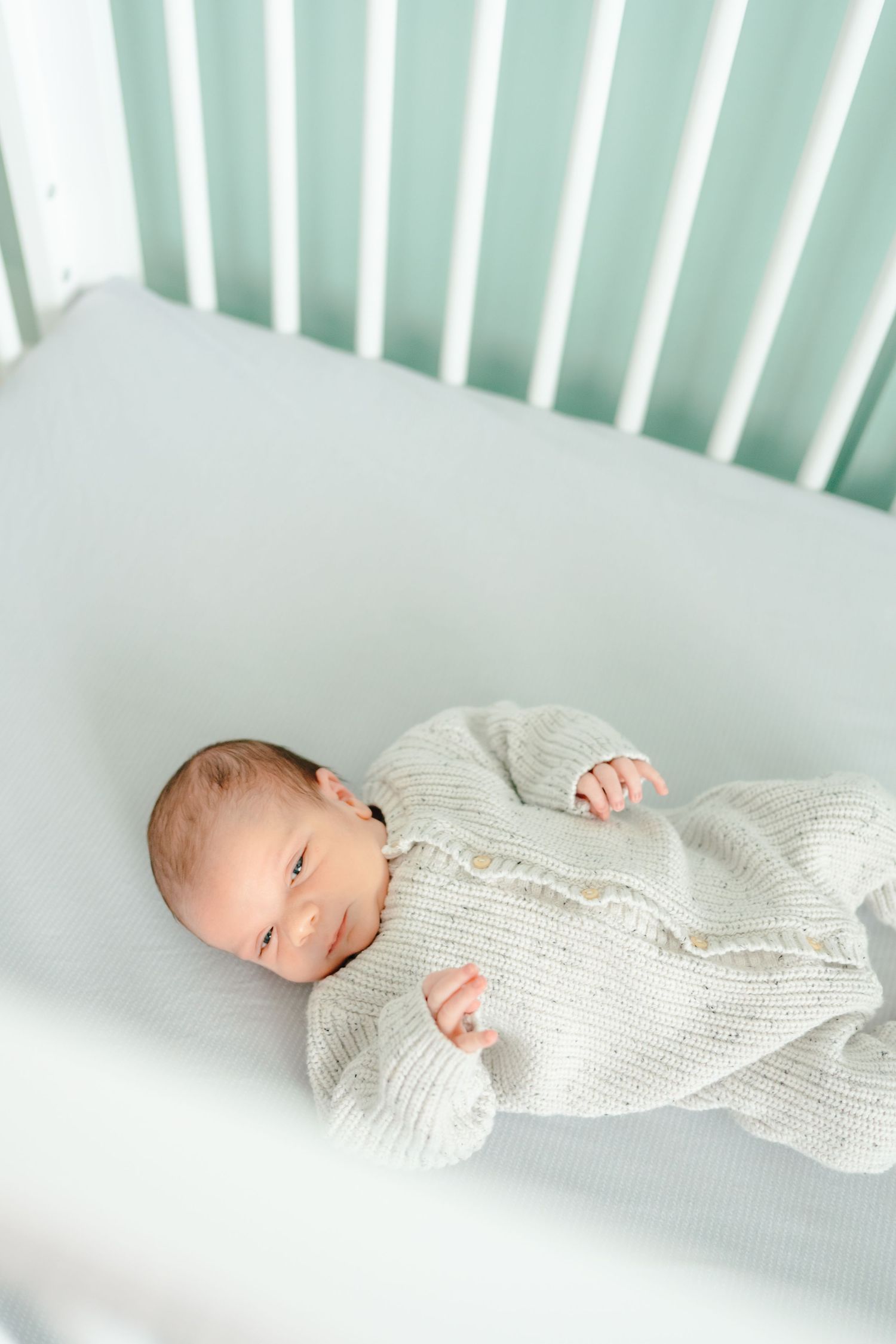 Baby in white knit outfit rests peacefully in white crib with mint green backdrop.