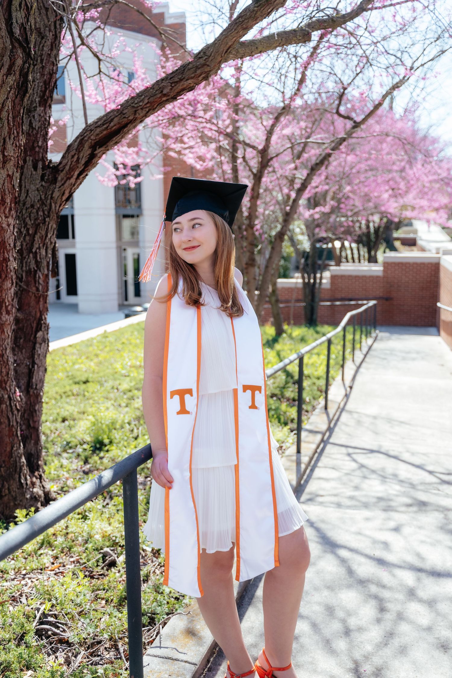 Graduate in cap and orange stole poses under blooming cherry trees on campus path.