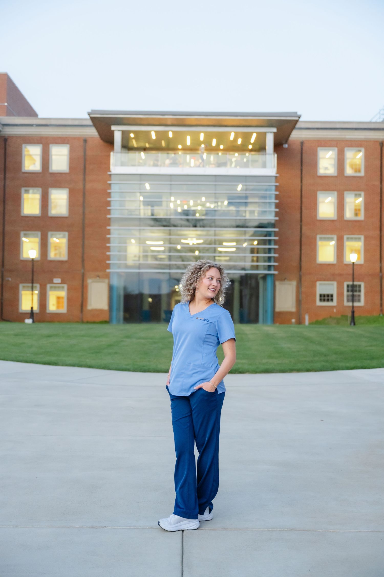 A female nurse in blue scrubs stands confidently outside a modern brick medical building at dusk.