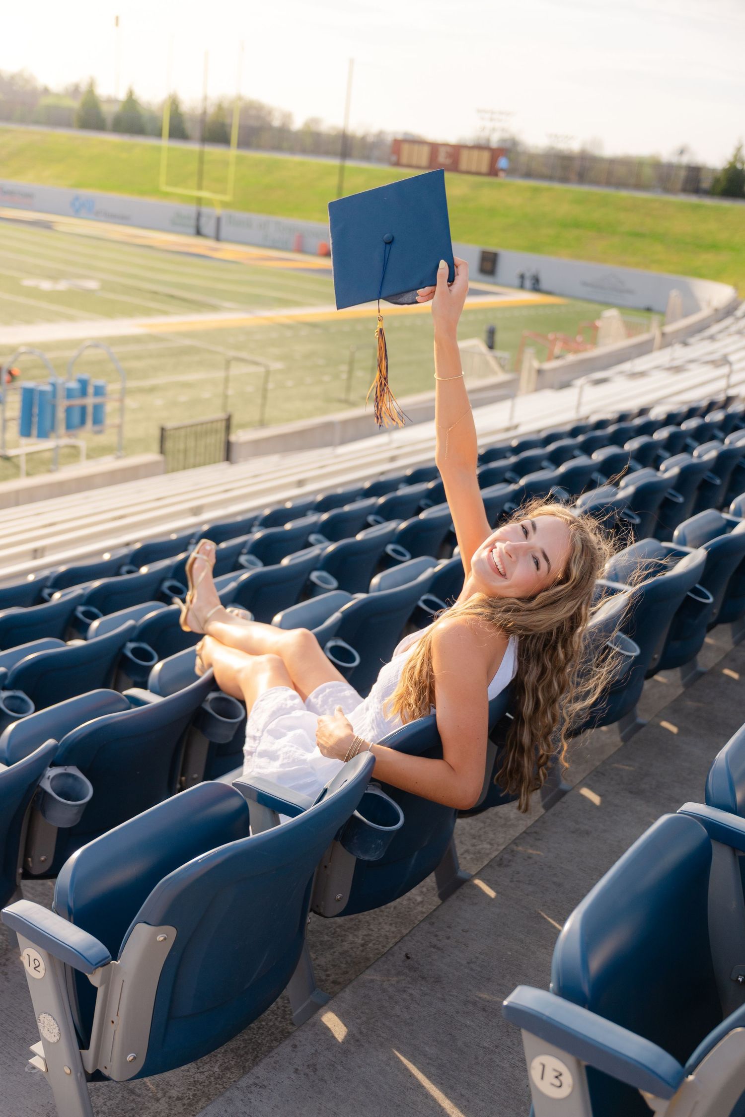 Smiling graduate raises blue cap in stadium bleachers, celebrating her graduation in a white dress.