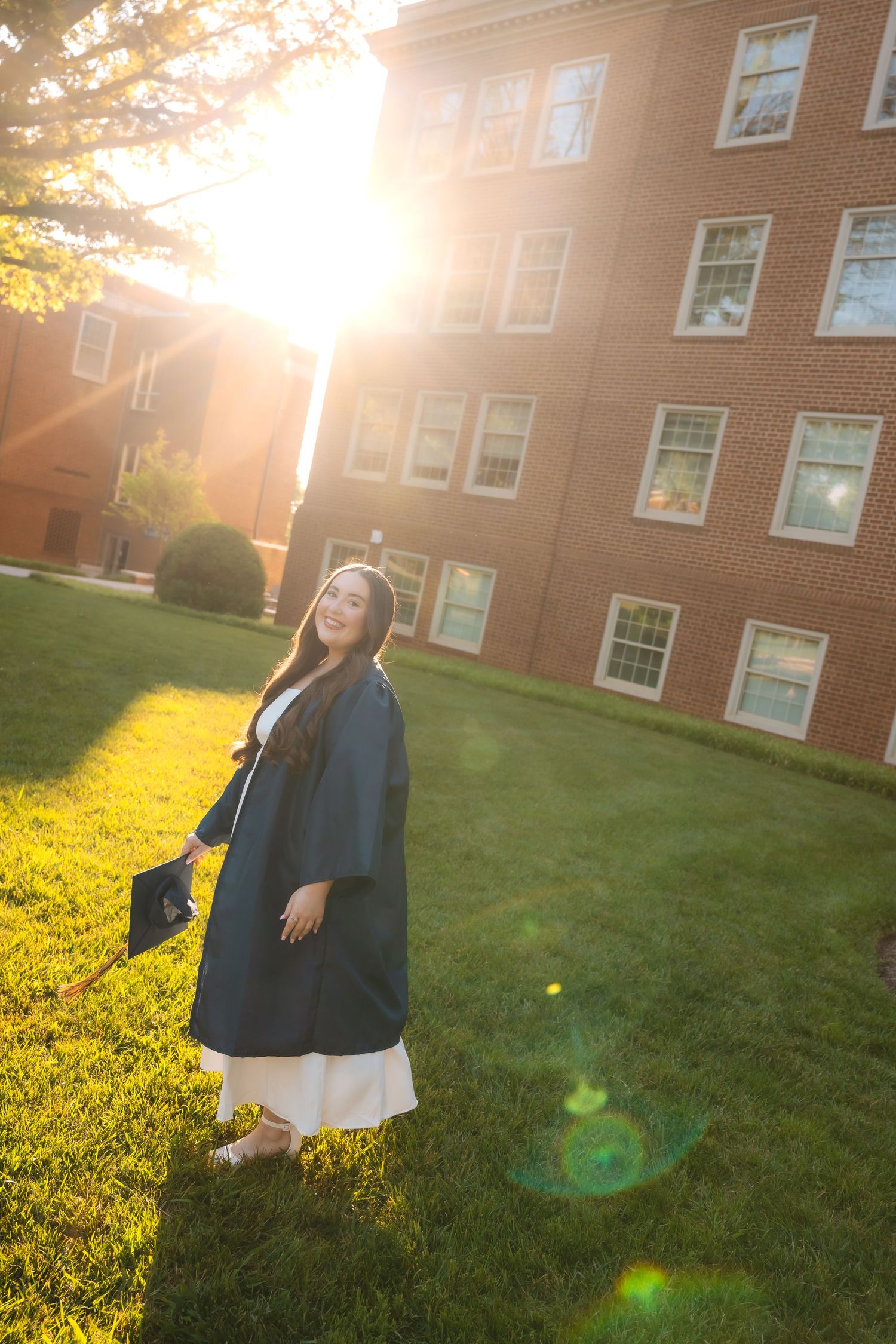 A young woman in graduation cap and gown stands on green campus lawn with golden sunlight flaring behind a brick building.