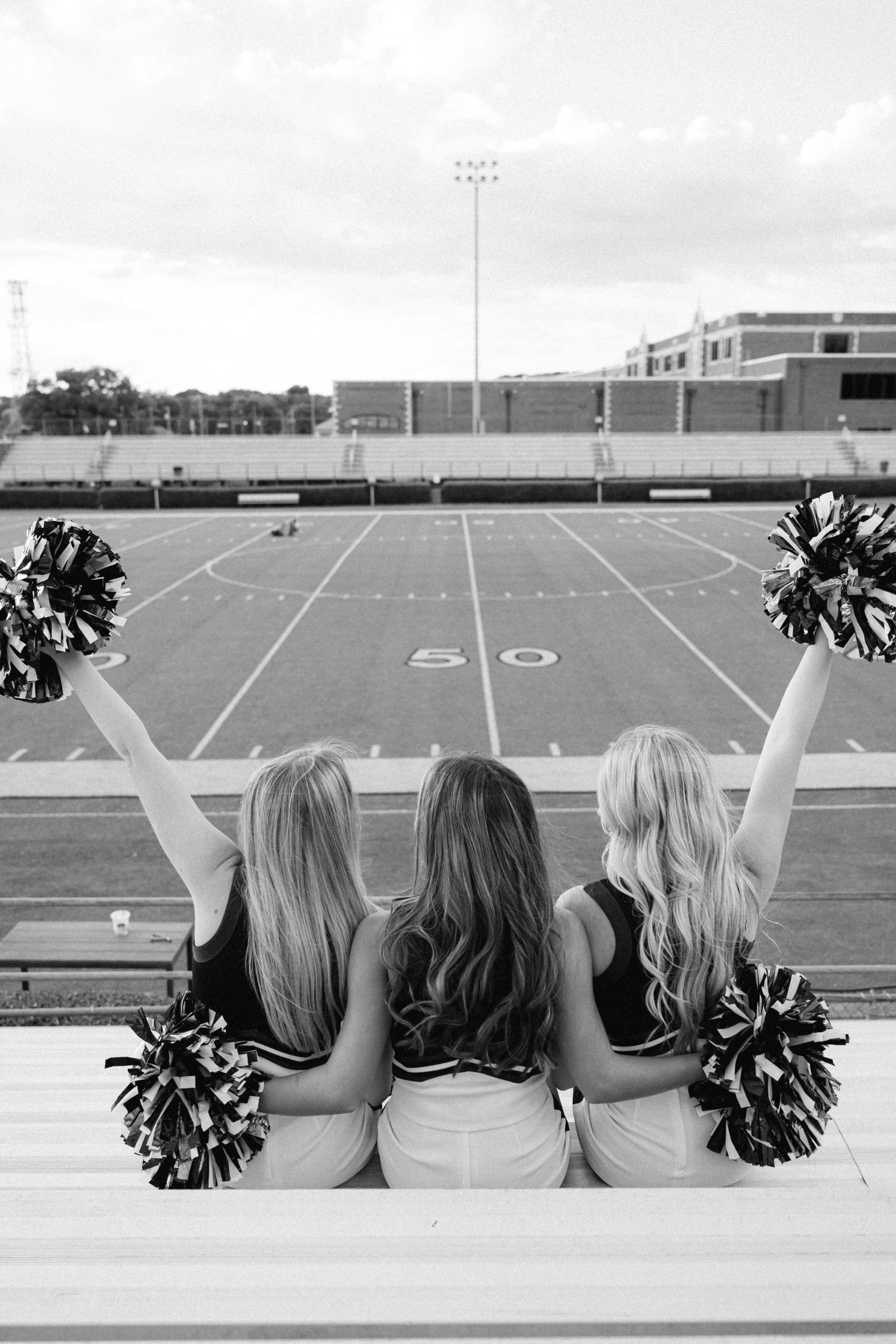 Three cheerleaders with pom-poms sit facing a football field at the 50-yard line in black and white photo.