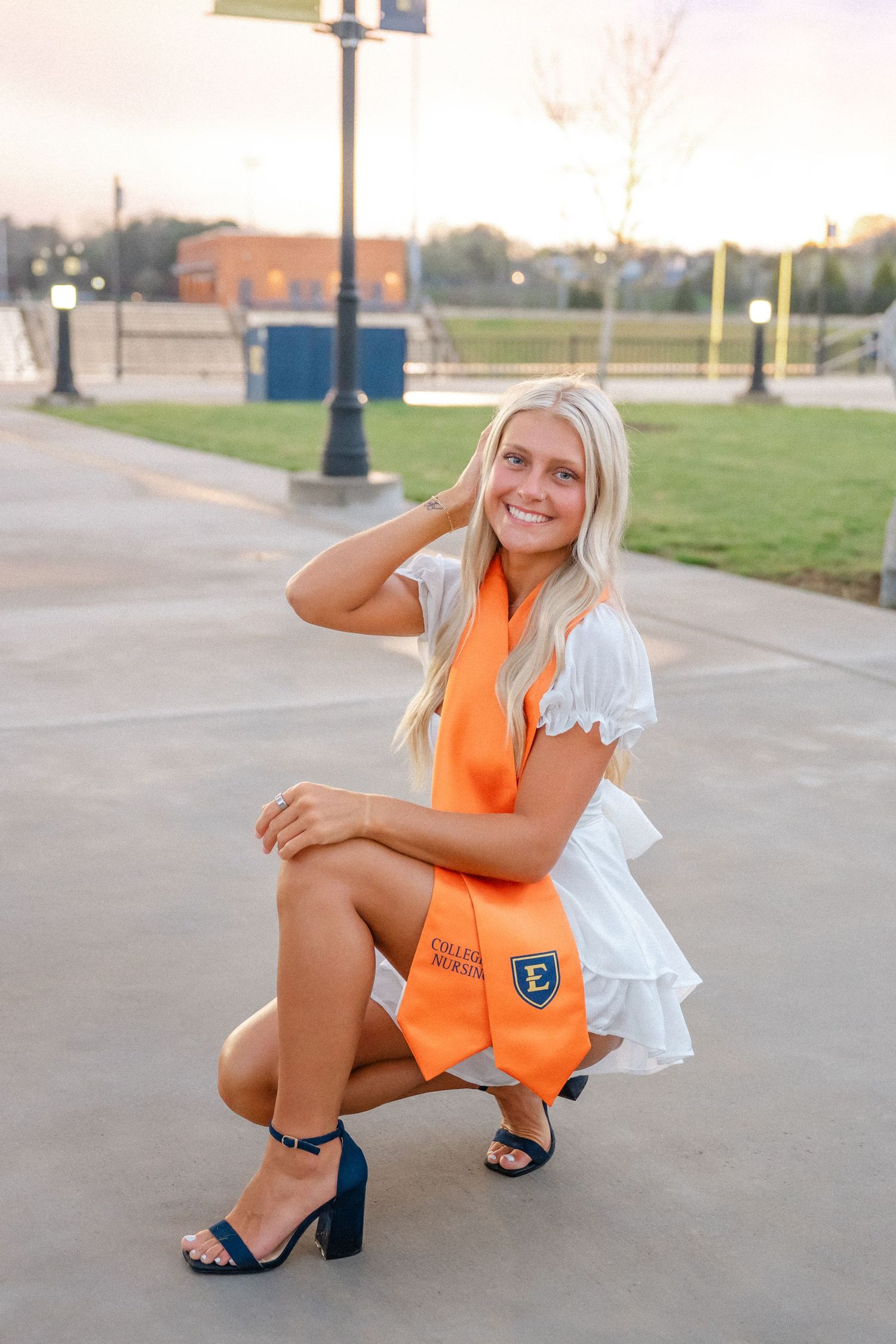 Smiling blonde woman in white dress and orange graduation stole crouching outdoors on campus at sunset.