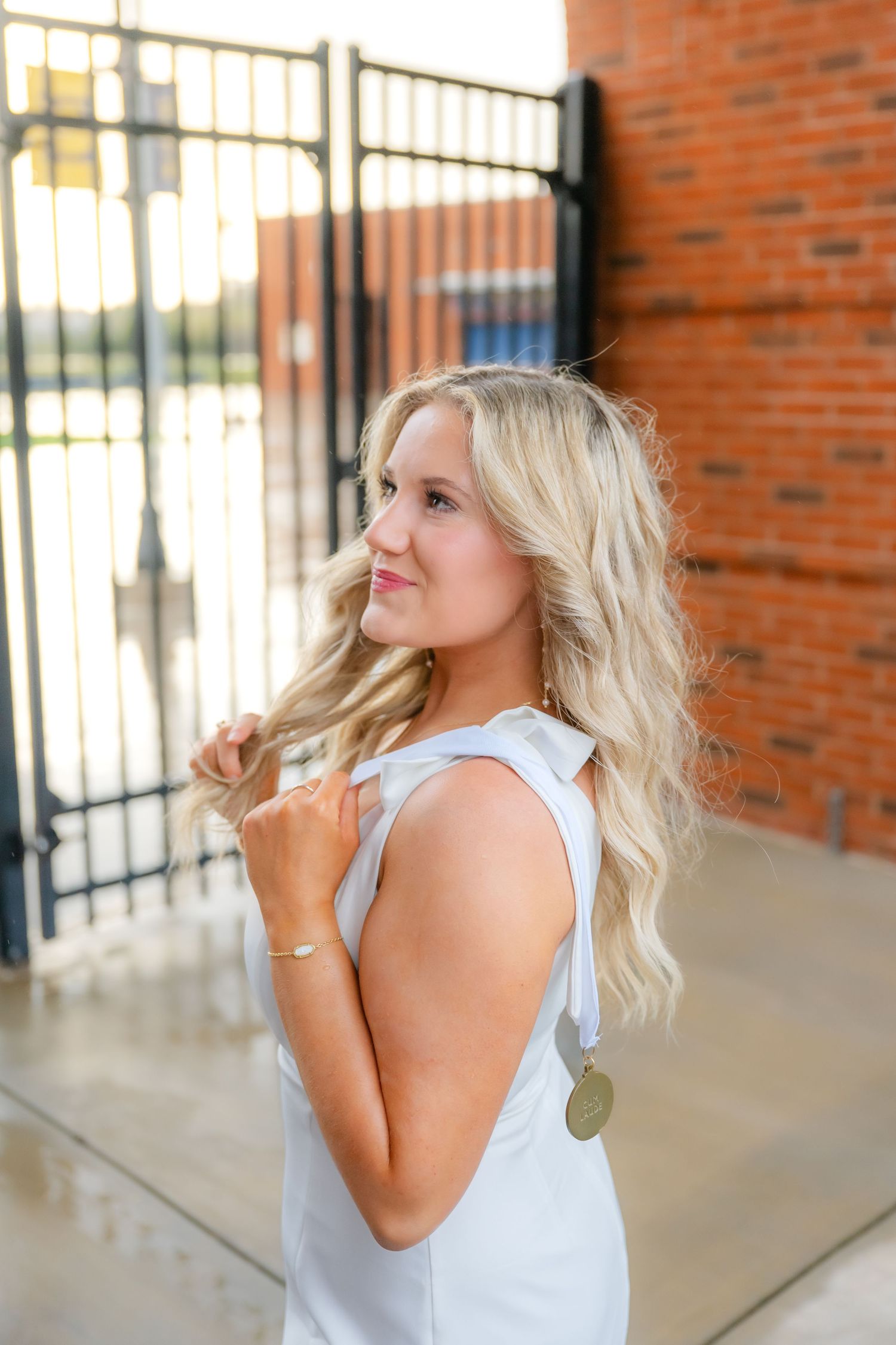 Blonde woman in white sleeveless dress poses outdoors near brick wall and iron gate, twirling her hair.