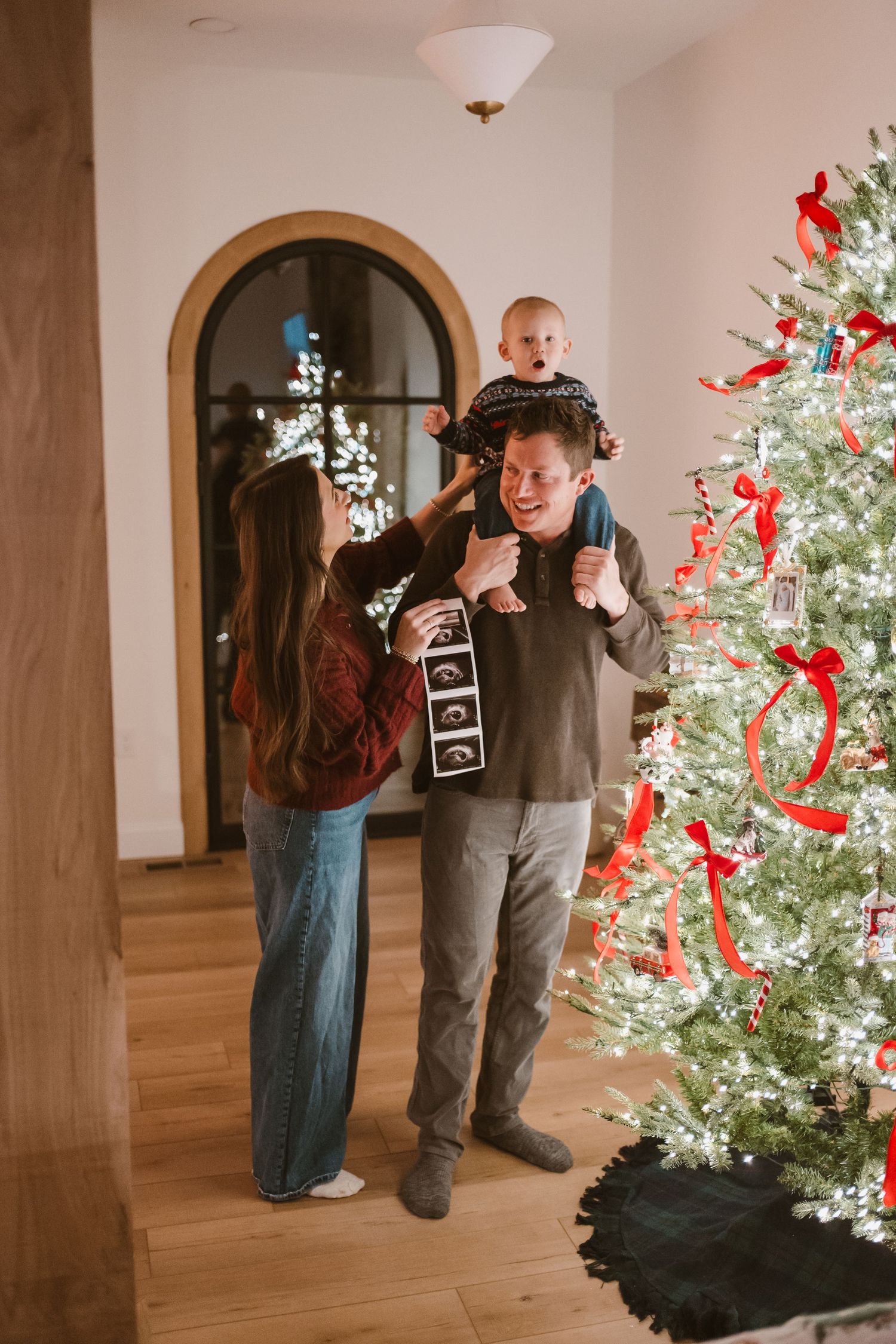 Family sharing pregnancy announcement with ultrasound strip by decorated Christmas tree in living room.