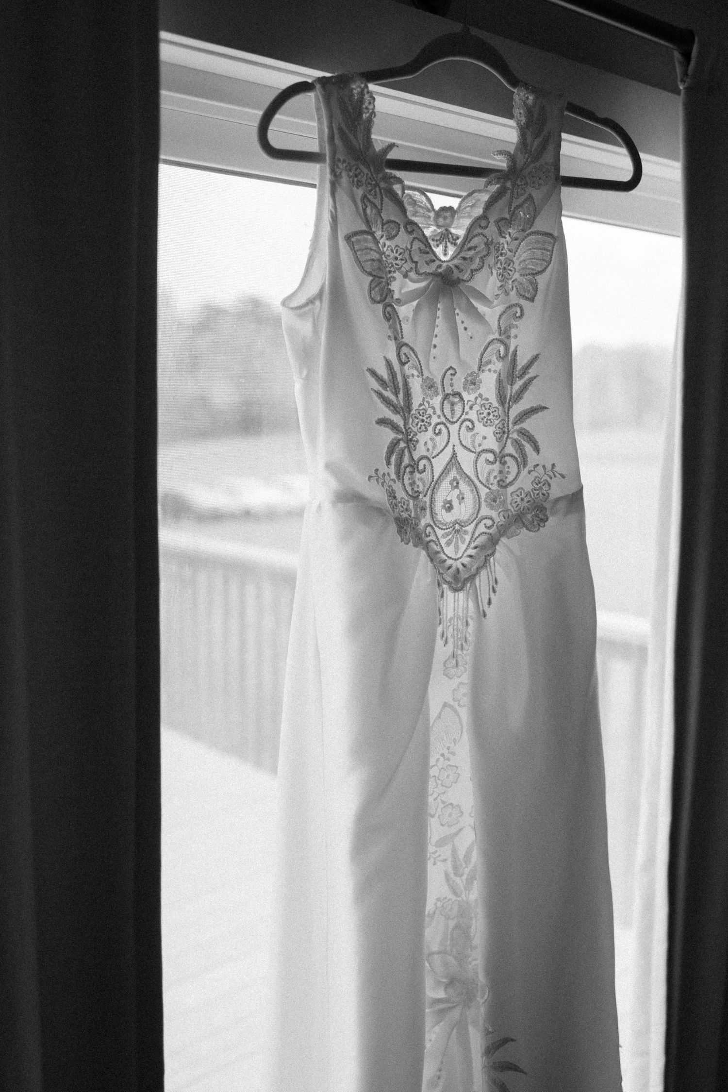 Delicate white wedding dress with lace detailing hanging by window in black and white.