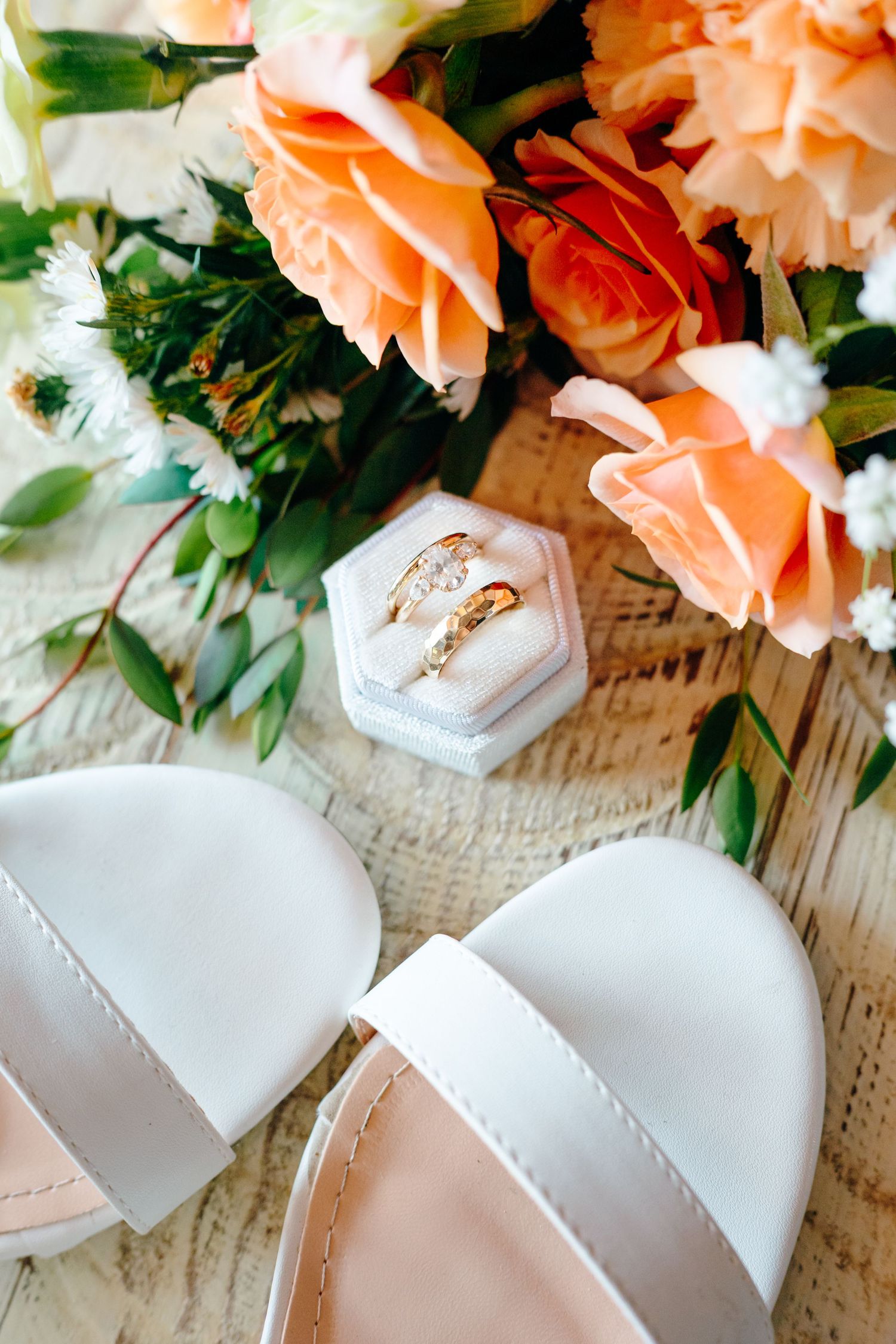 Wedding rings in hexagonal box with peach roses and white sandals on textured surface.